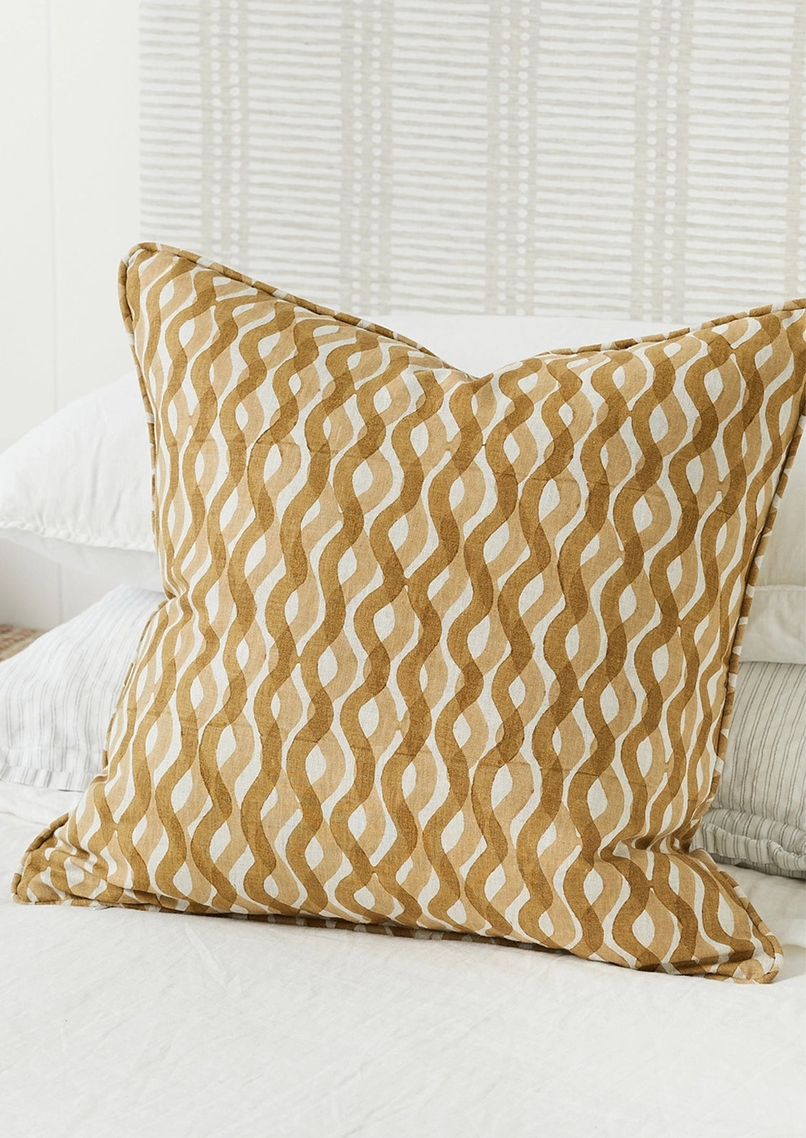 A square decorative throw pillow with a wavy, interlocking geometric pattern in warm tan and off-white, styled on a white bed.