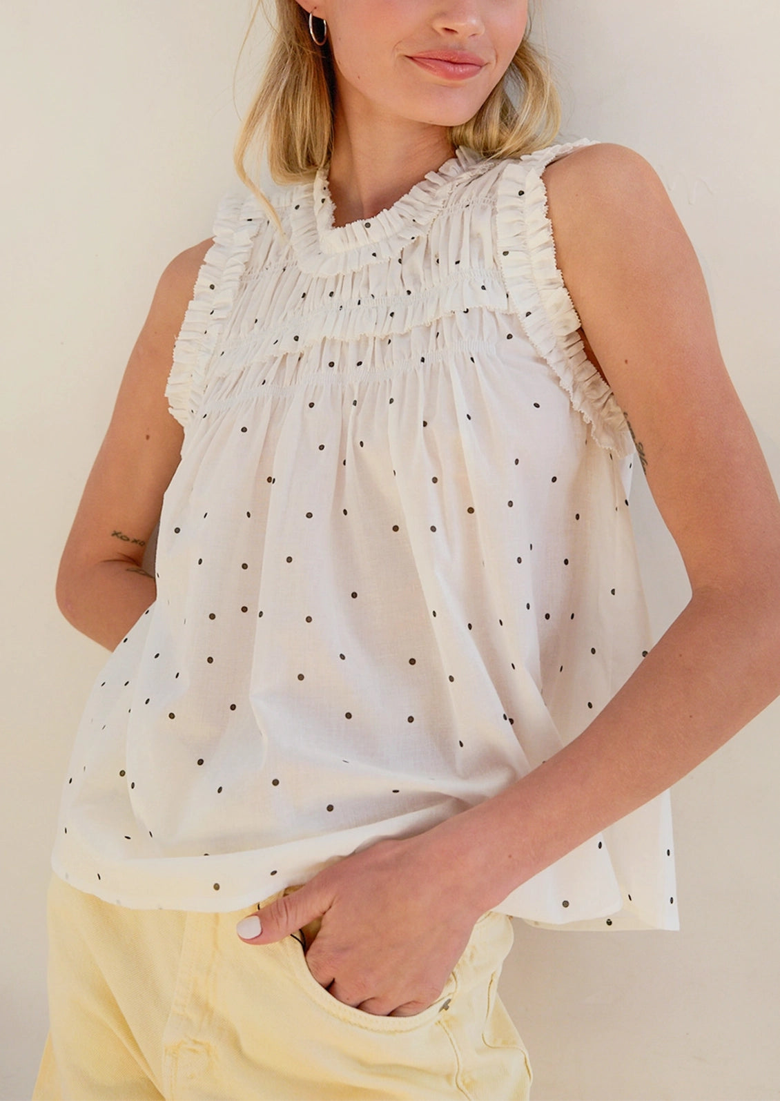 A woman wearing a sleeveless white top with small black polka dots and ruffled trim, paired with pale yellow pants, against a plain light beige background.