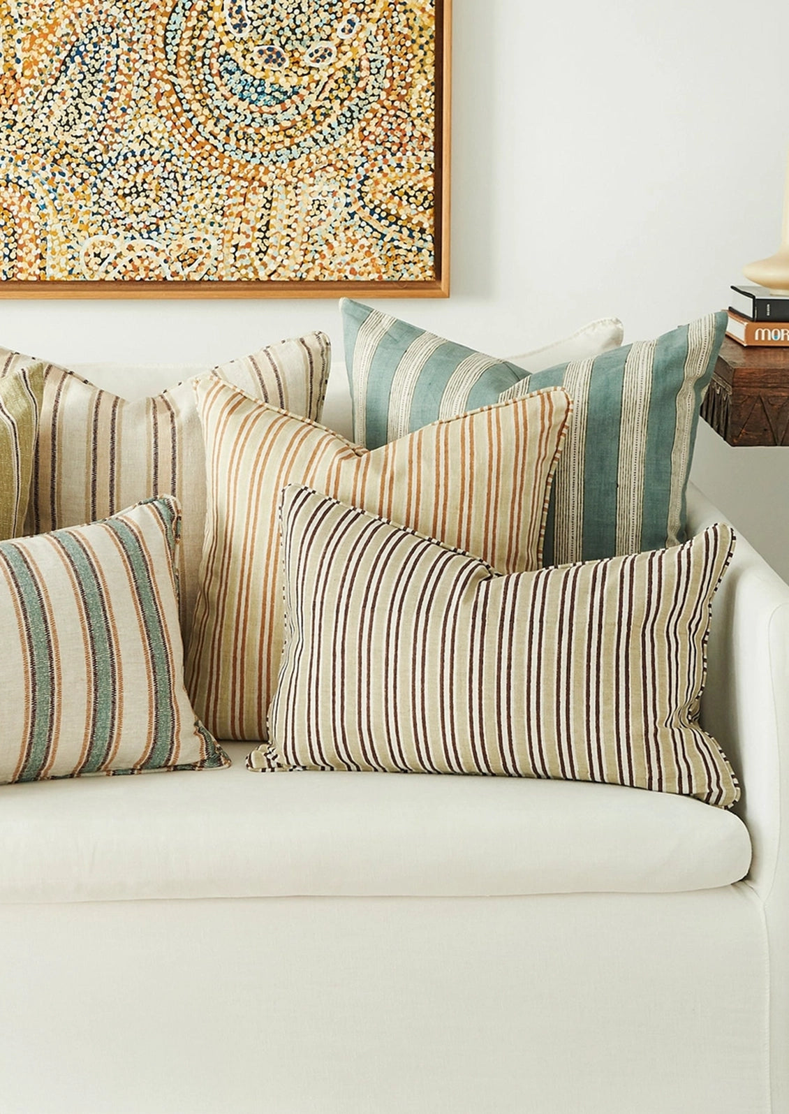 A set of striped throw pillows in beige, cream, and teal tones arranged on a white sofa, with a framed abstract painting and a side table visible in the background.