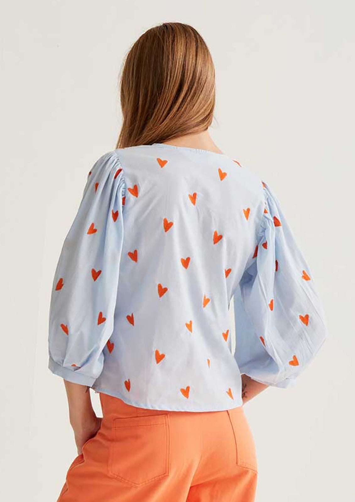 Woman wearing a light blue blouse with red heart patterns and orange pants on a white background