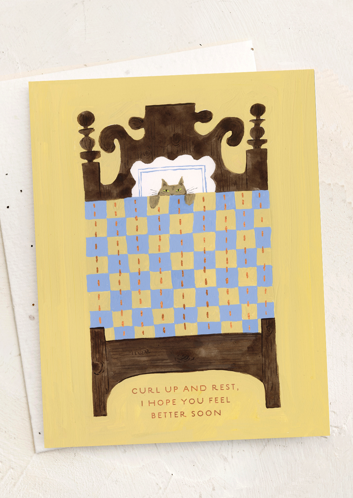 Greeting card with a cat on a checkered bed, text 'curl up and rest, I hope you feel better soon' on a yellow background.