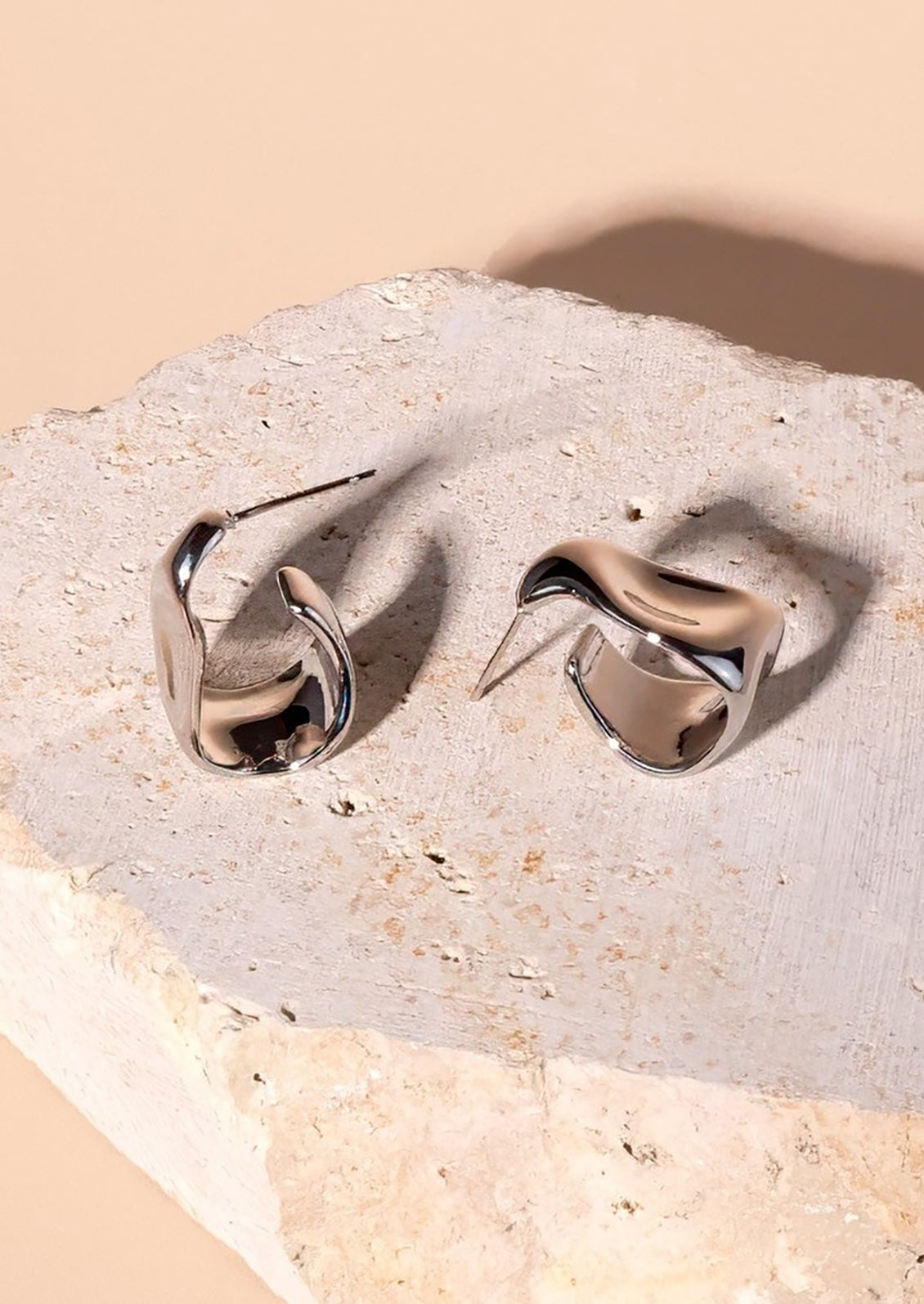 A pair of asymmetrical silver hoop earrings with hammered wavy texture.
