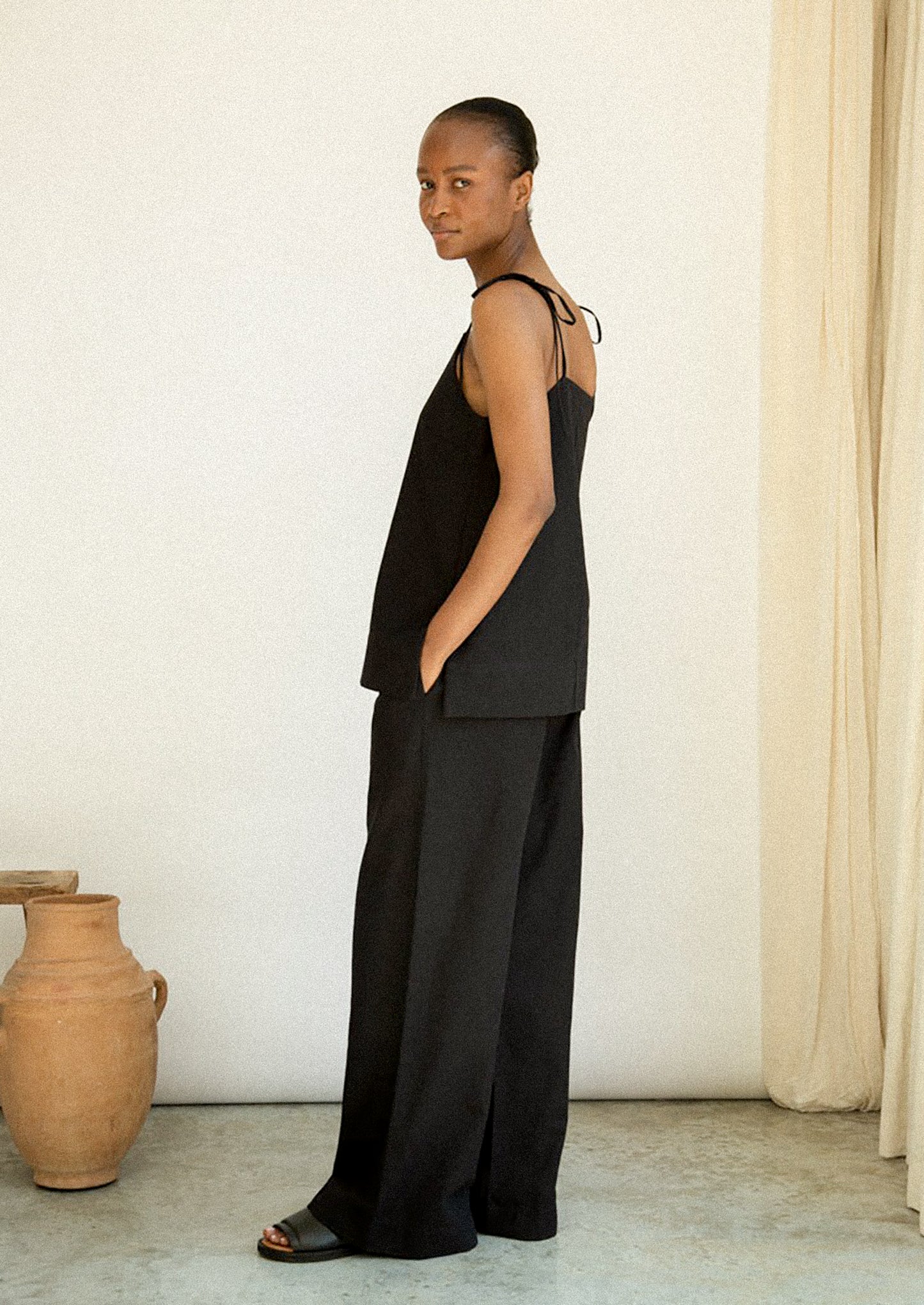 Woman wearing a black sleeveless top and wide-leg pants standing against a white wall with a beige curtain.