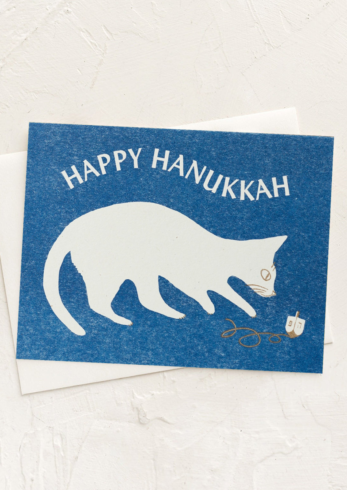 A risograph printed card in blue with cat playing with dreidel, text reads "Happy Hanukkah".
