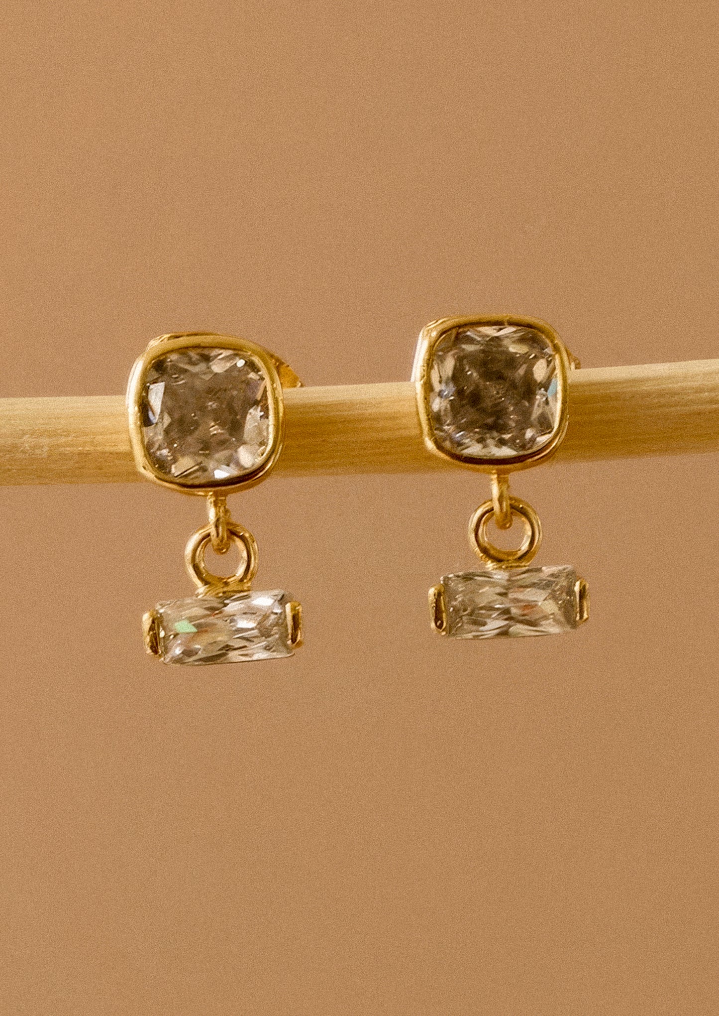 A pair of gold drop earrings with square cushion-cut clear crystals on the top and smaller rectangular clear crystals below, displayed on a light wooden stick against a warm tan background.
