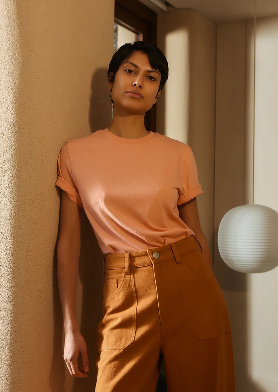 Person wearing a peach t-shirt and brown pants standing against a beige wall.