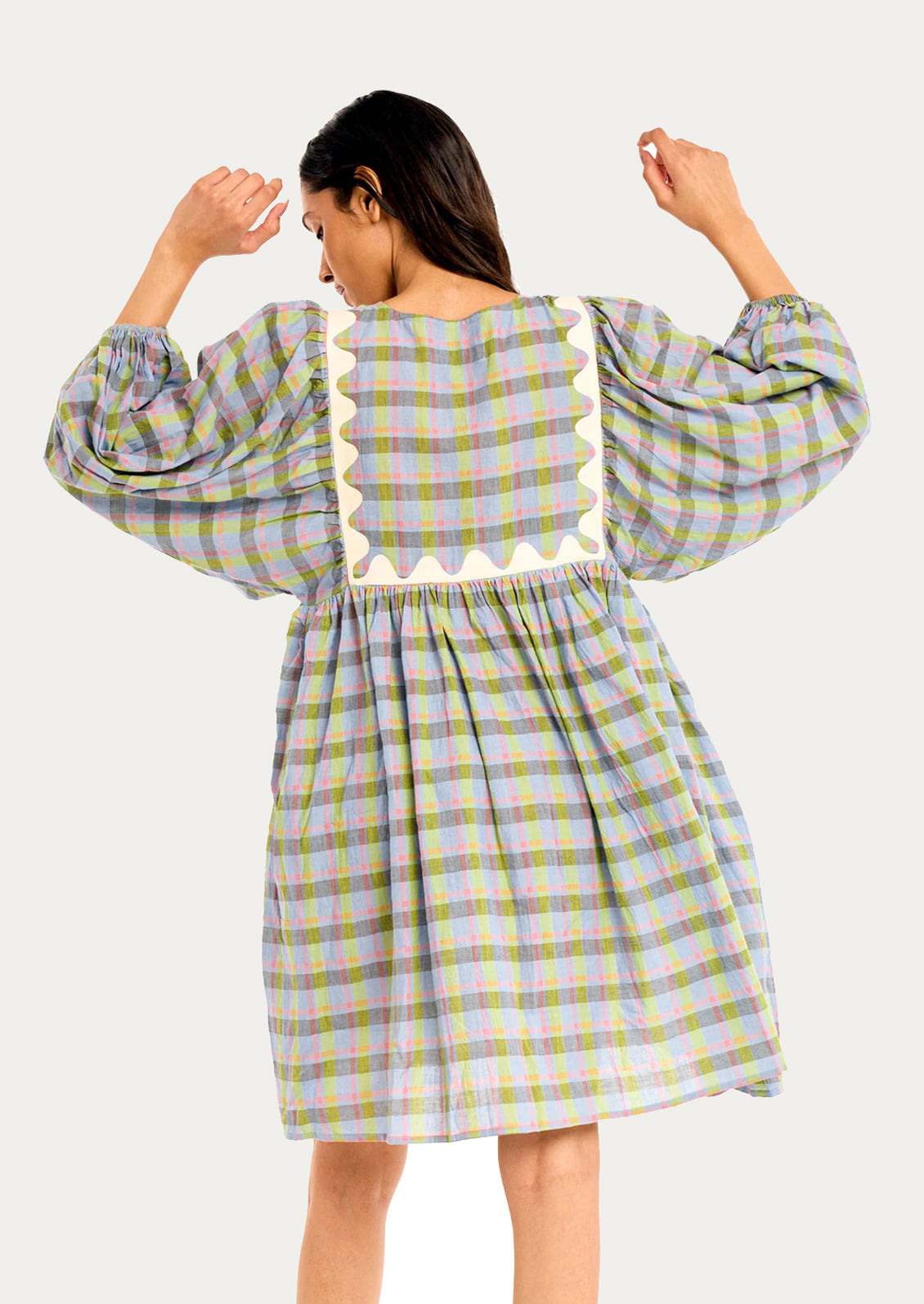 A woman shown from the back wearing a long-sleeve plaid dress with a cream scalloped trim along the back neckline, set against a solid light gray background.