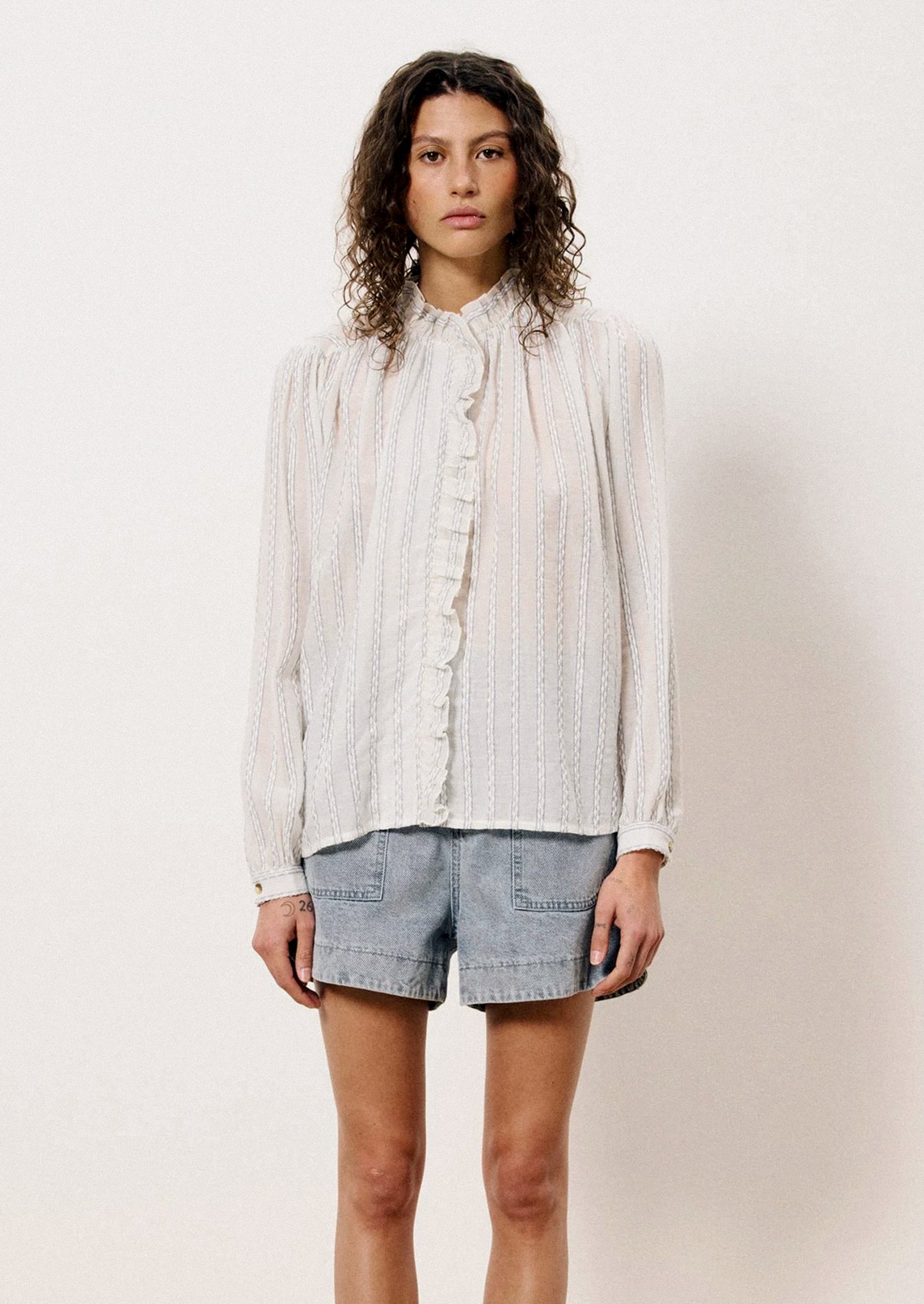 A woman wearing a long sleeve blouse with ruffle front placket and interwoven stripe detail throughout.
