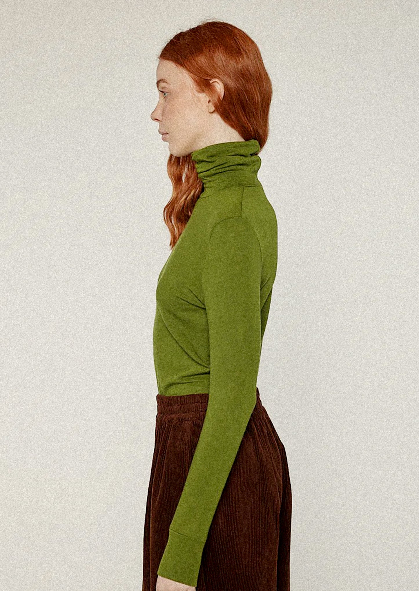 Person wearing a green turtleneck sweater and brown pants against a white background