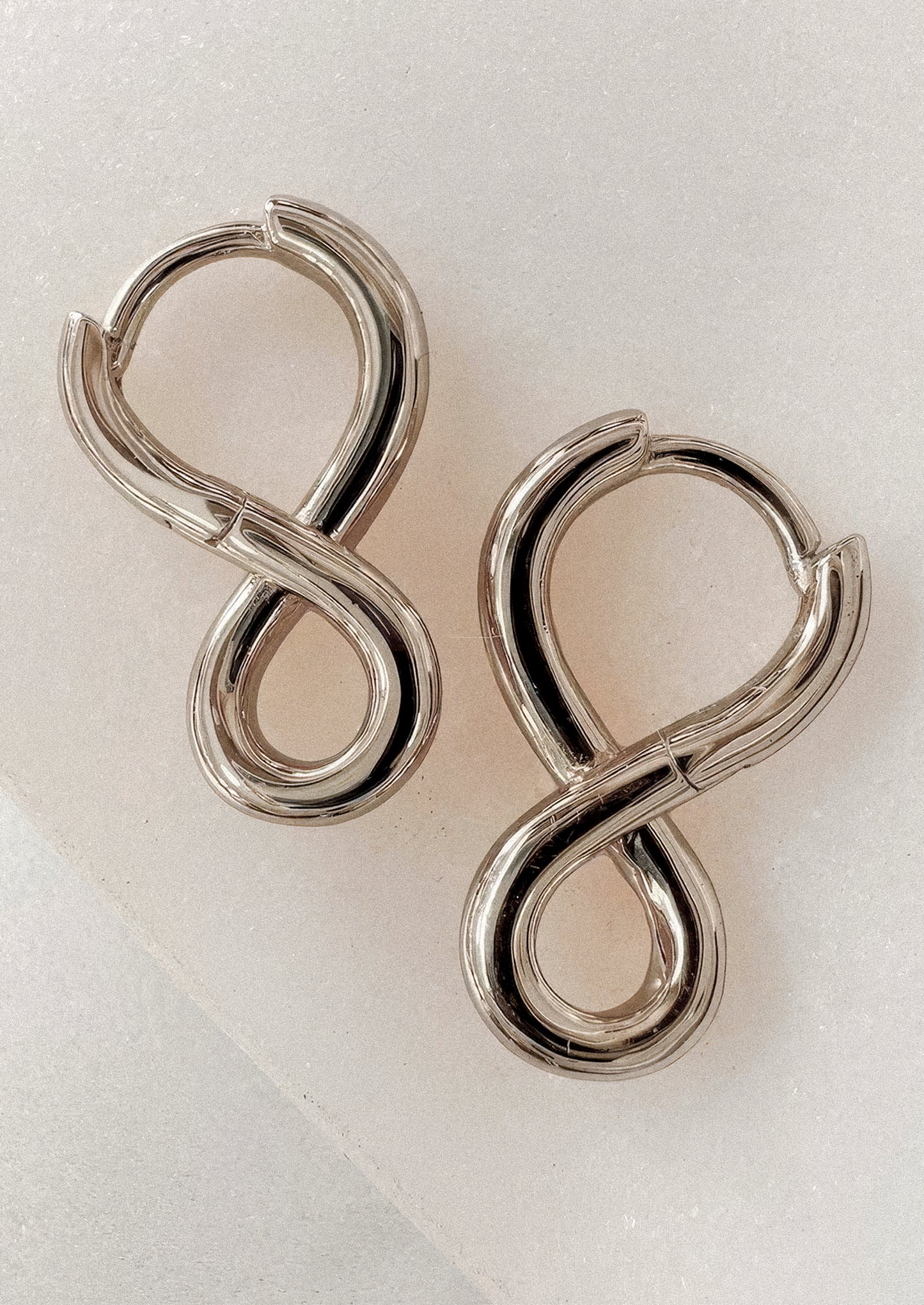 A pair of polished silver infinity-shaped hoop earrings on a textured white background.