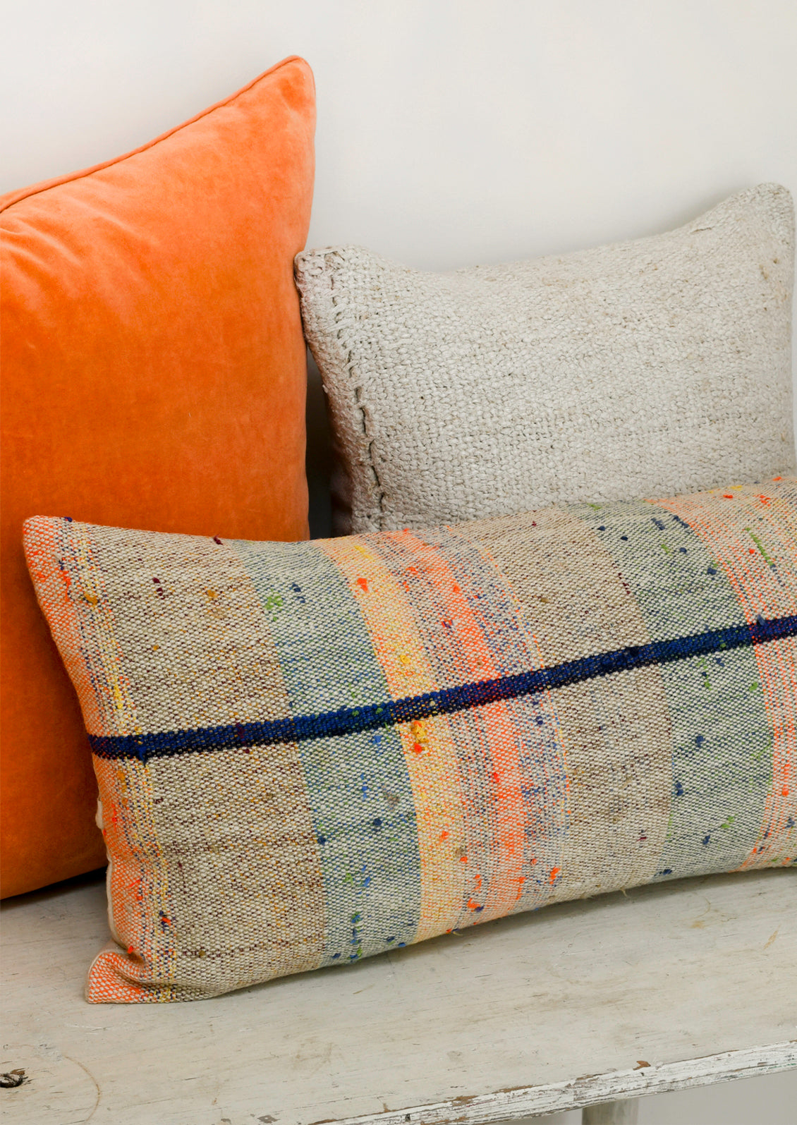 A rectangular 12" x 24" mottled kilim pillow with various colored striped throughout sitting on a table with two other pillows.