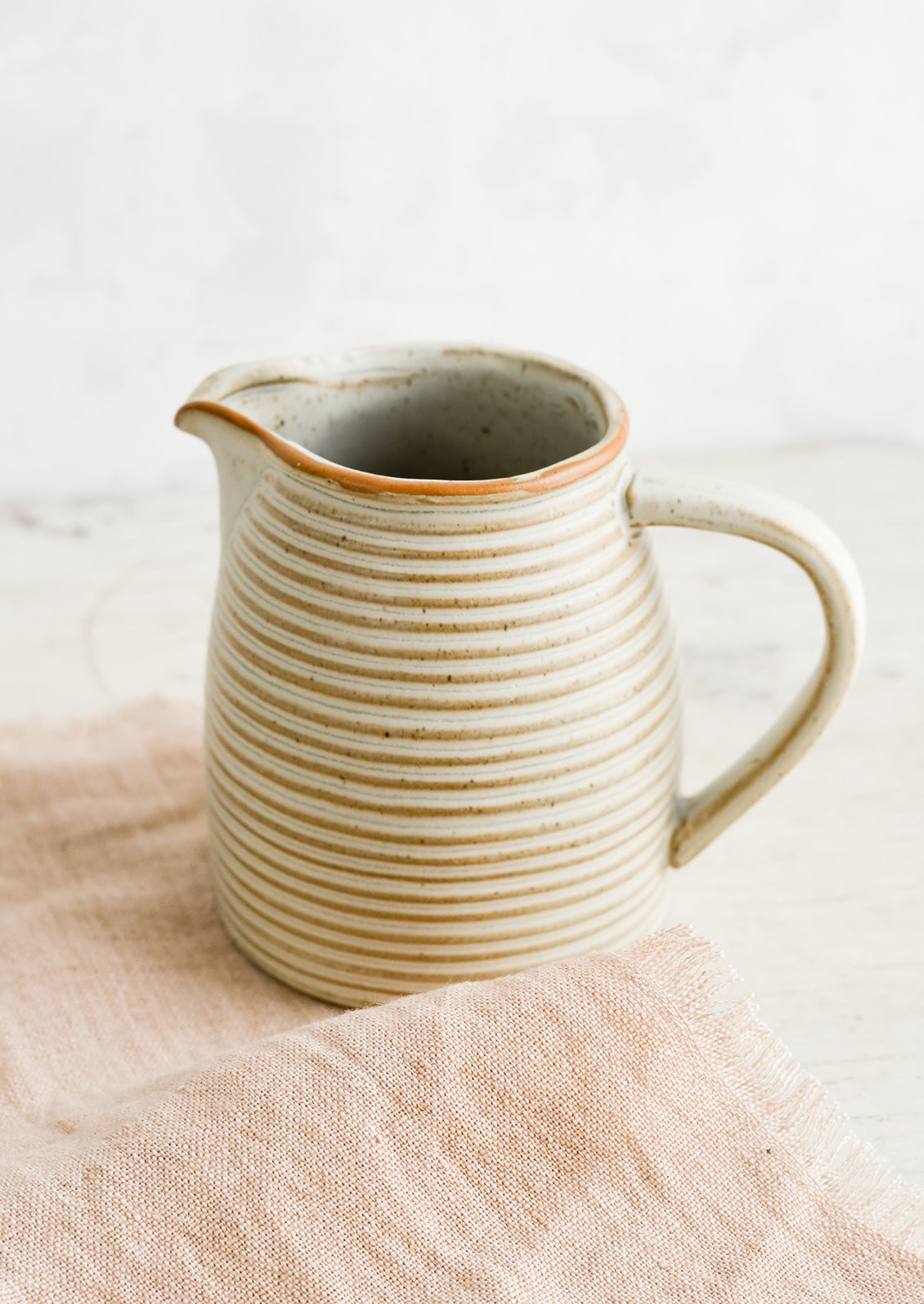 A tan ceramic pitcher with striped texture on top of pink linen.