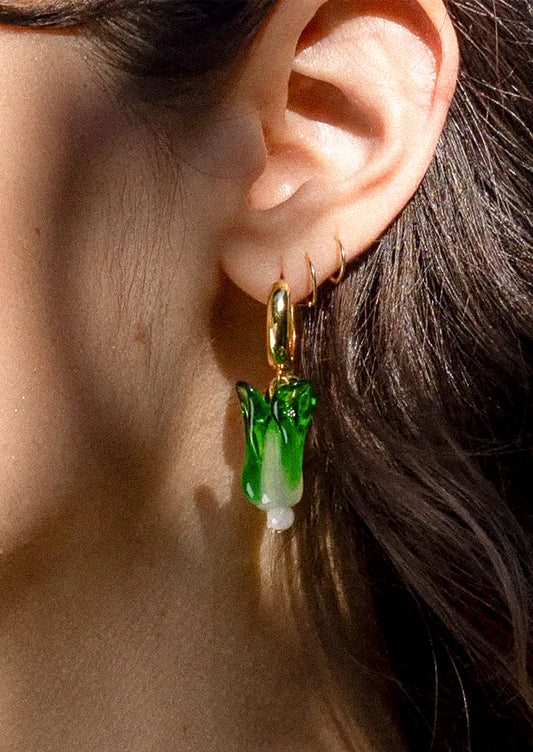 A close-up of a person's ear wearing a dangling earring with a gold-tone hook and a green-and-white translucent vegetable-shaped charm.