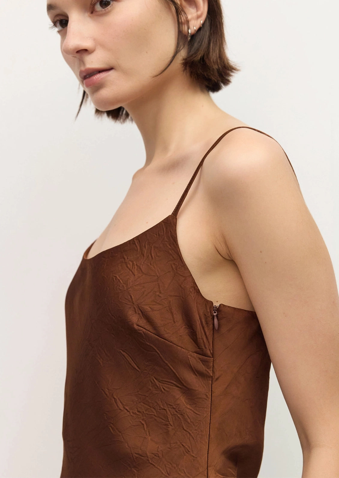 A close-up side view of a woman wearing a brown crinkled-texture spaghetti-strap camisole top against a plain light background.