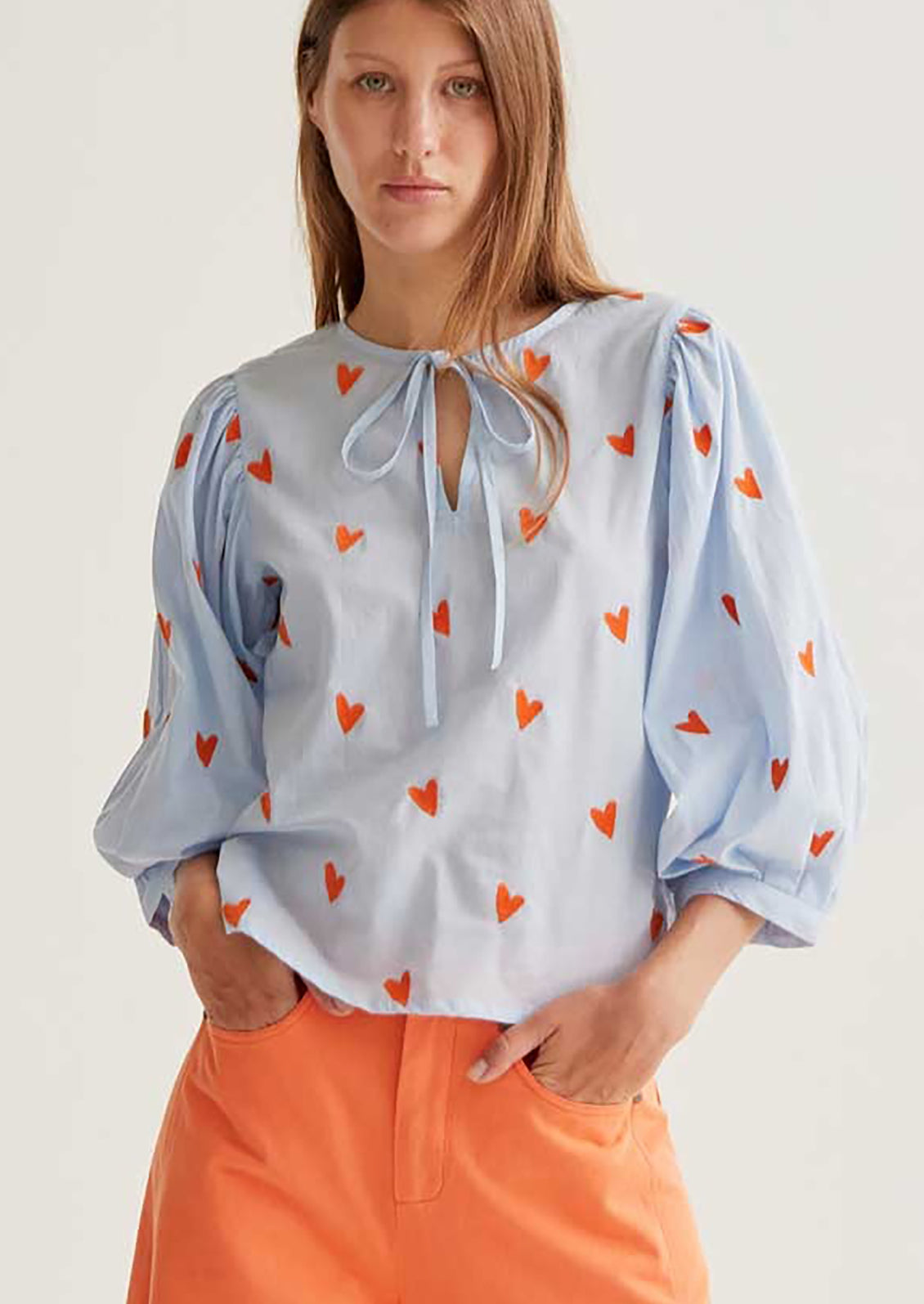 Woman wearing a light blue blouse with orange heart patterns and orange pants against a white background