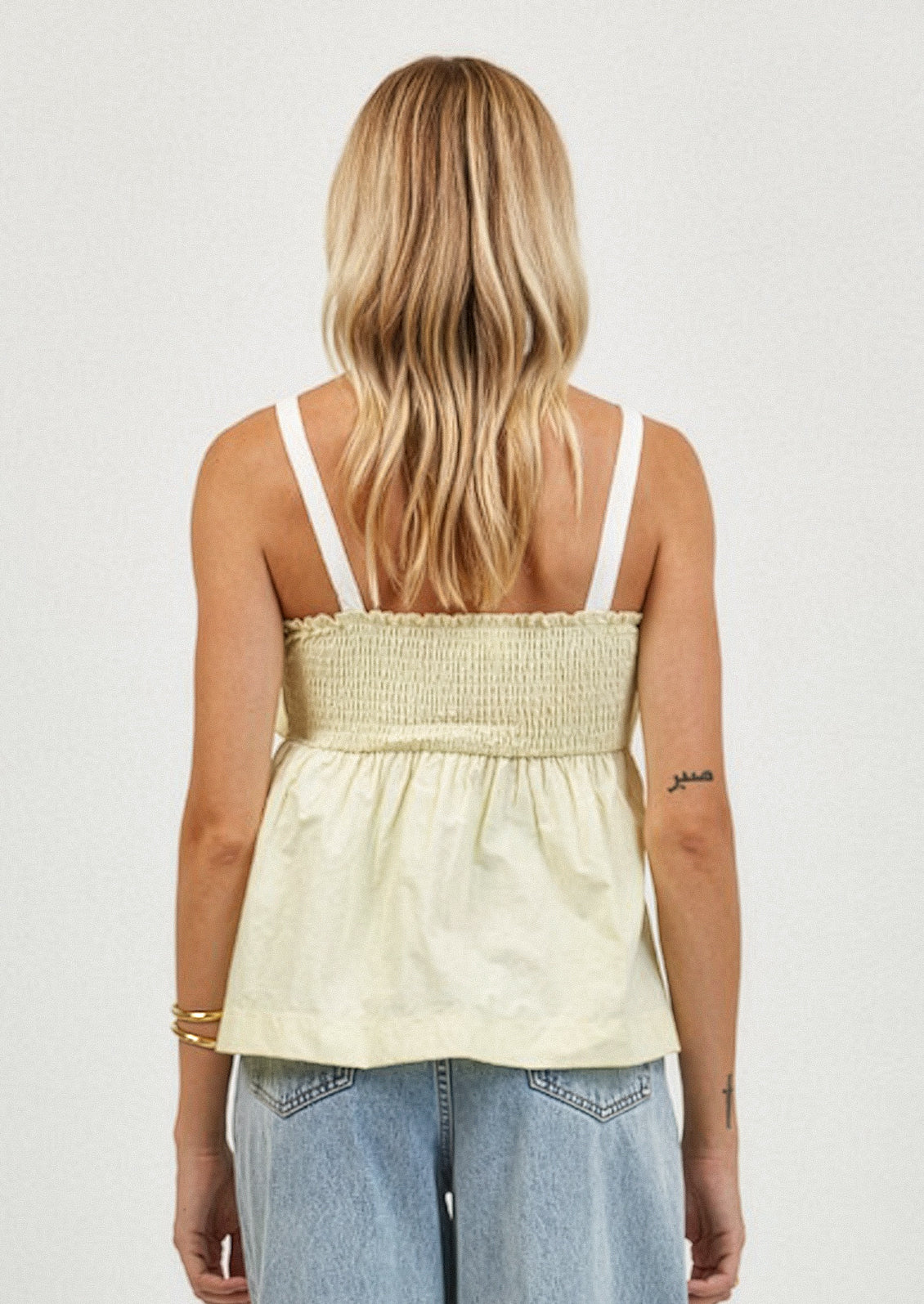 Woman wearing a light-colored top with thin straps and denim pants on a plain background