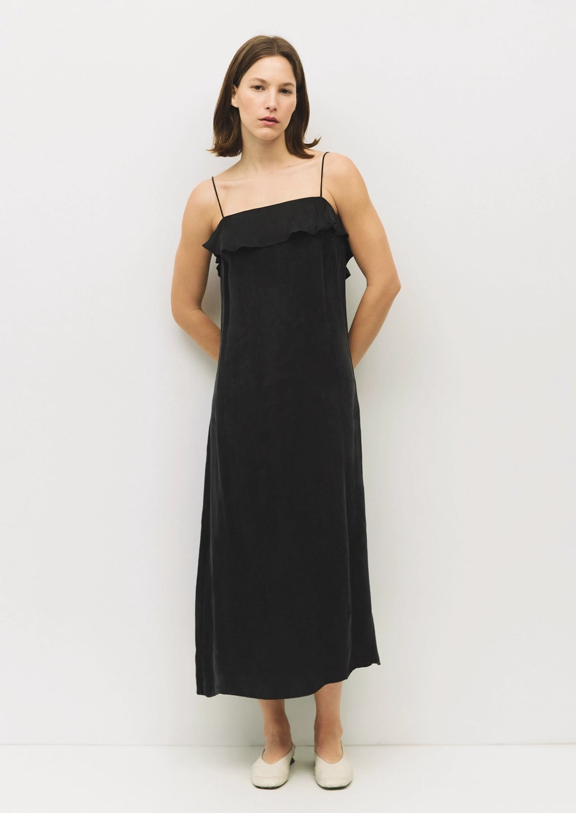 A woman modeling a black midi-length spaghetti-strap dress with a ruffled neckline, paired with off-white flat shoes, set against a plain white studio background.