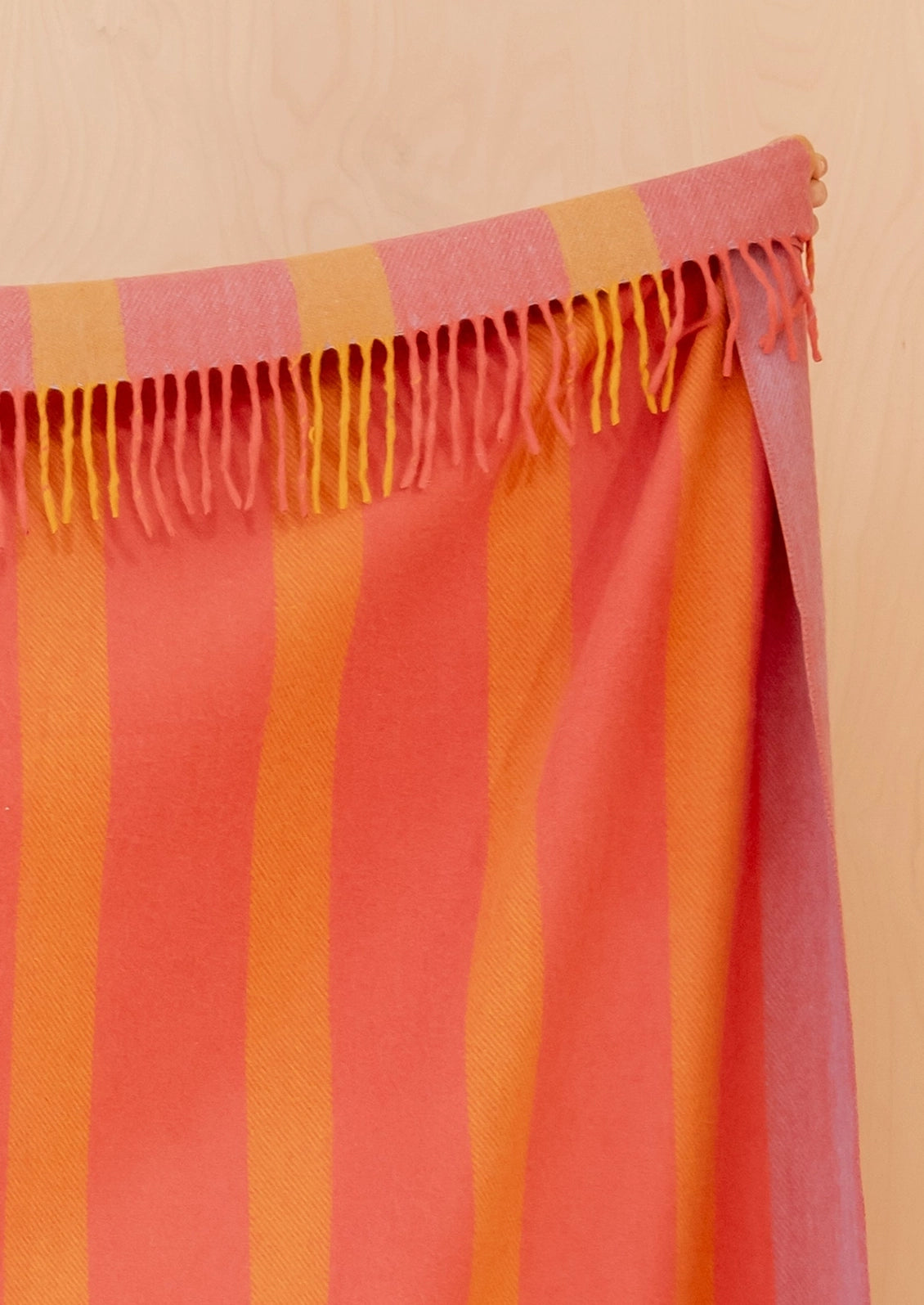 A close-up of a striped blanket with fringe along the top edge, shown against a light wood background. The fabric features vertical stripes in coral pink, orange, and yellow.