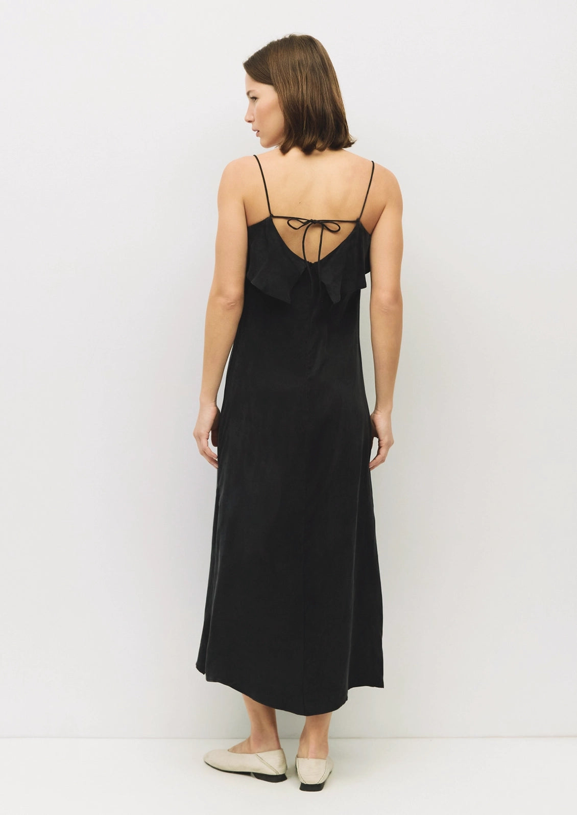 A woman wearing a black midi slip dress with thin straps and a ruffled tie-back neckline, shown from the back against a plain white studio background.