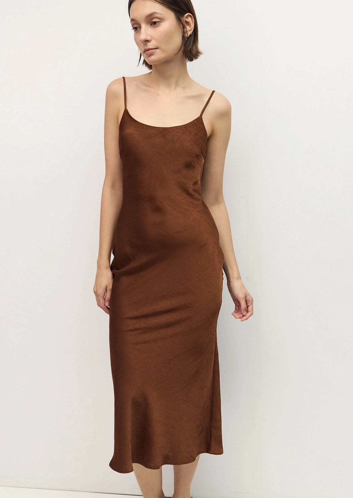 A woman modeling a brown satin slip dress with thin spaghetti straps and a scoop neckline, standing against a plain off-white wall.