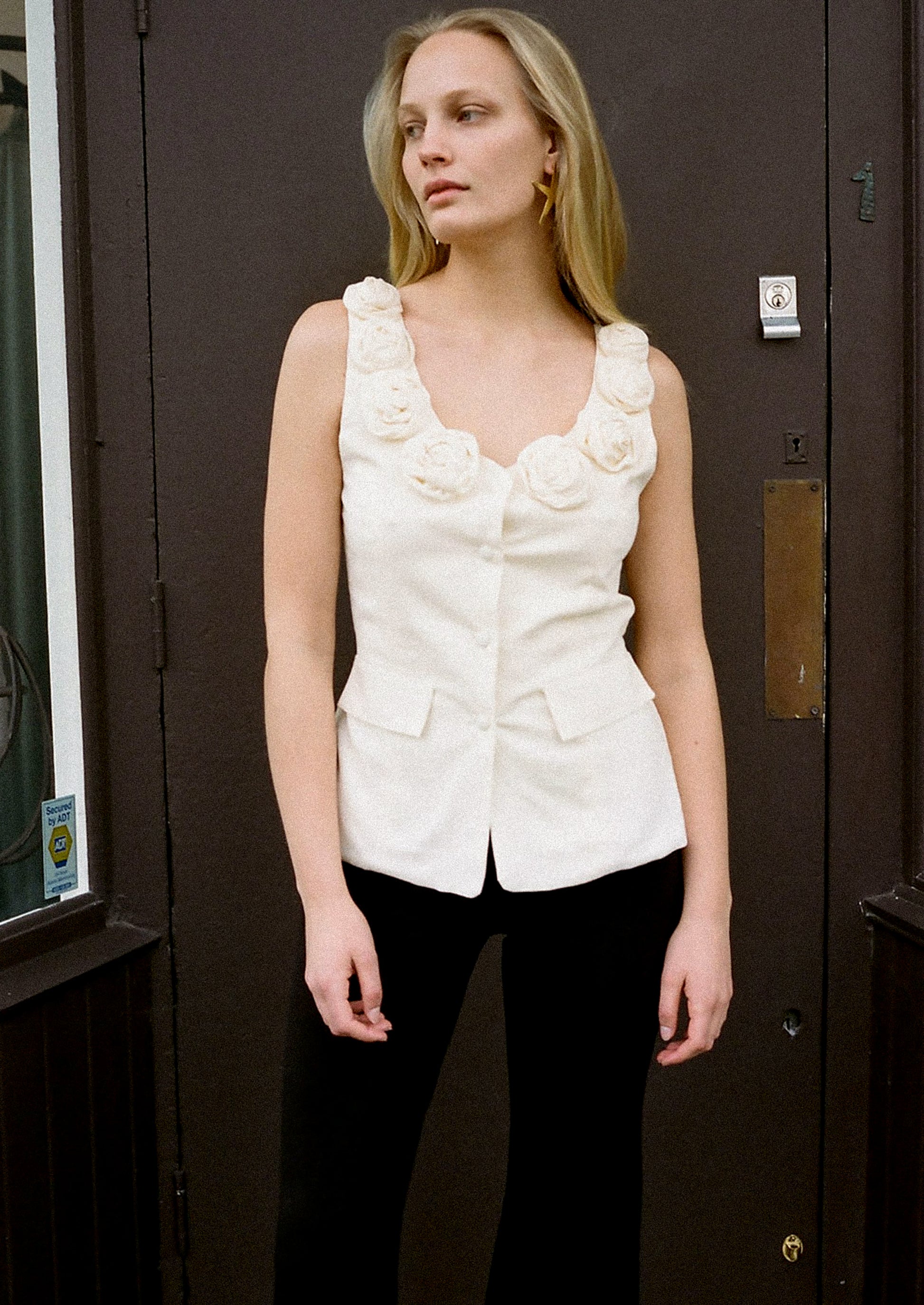 A woman wearing a cream u-neck blouse with dramatic rosette detail along neckline.