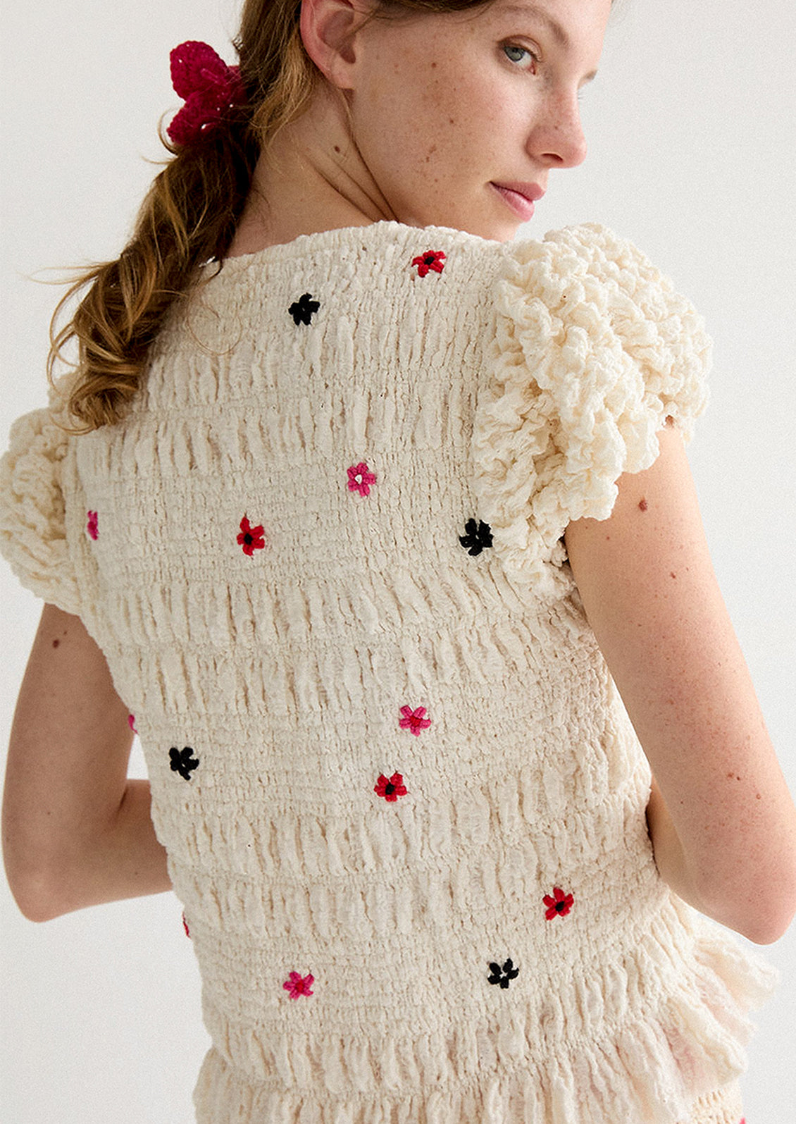 A woman wearing a smocked and ruffled cream top with red and pink embroidered flowers throughout.