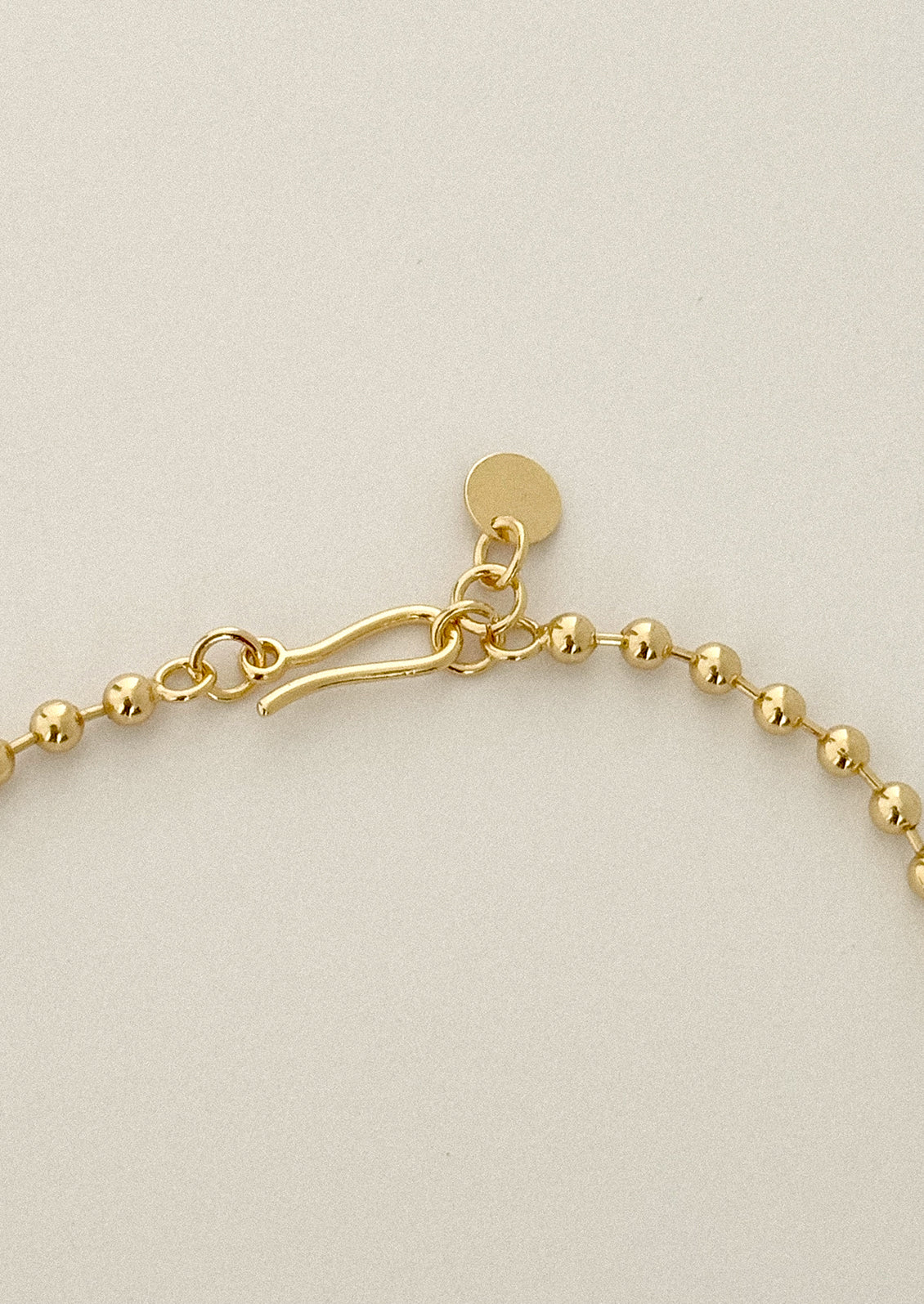 Close-up of a gold beaded chain with a toggle clasp and a small round tag, shown on a light neutral background.