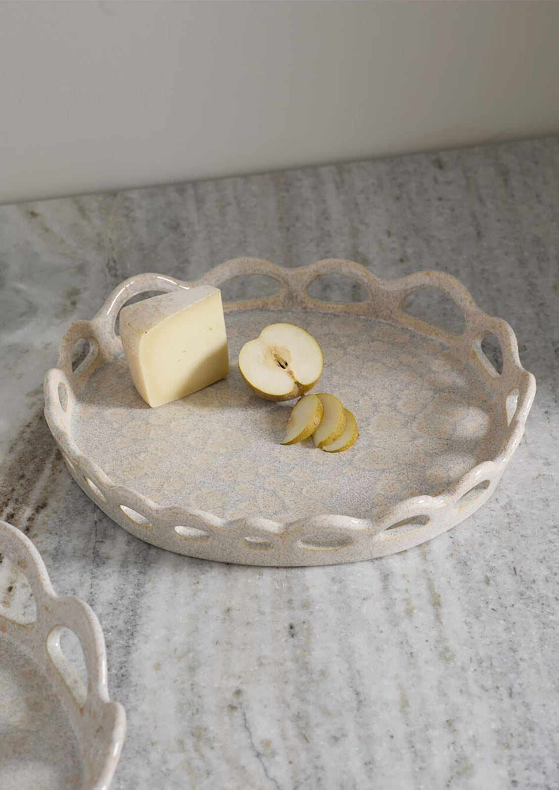 A round ceramic serving tray with a scalloped cutout edge, shown on a marble surface with cheese and apple slices as props.