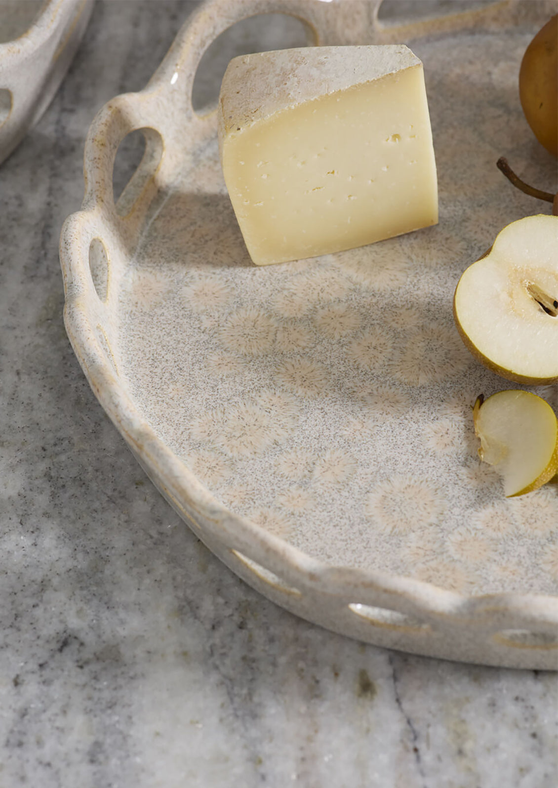 A close-up of a round speckled beige ceramic serving tray with scalloped handles, styled with a wedge of cheese, a sliced pear, and a whole pear on a gray marble surface.