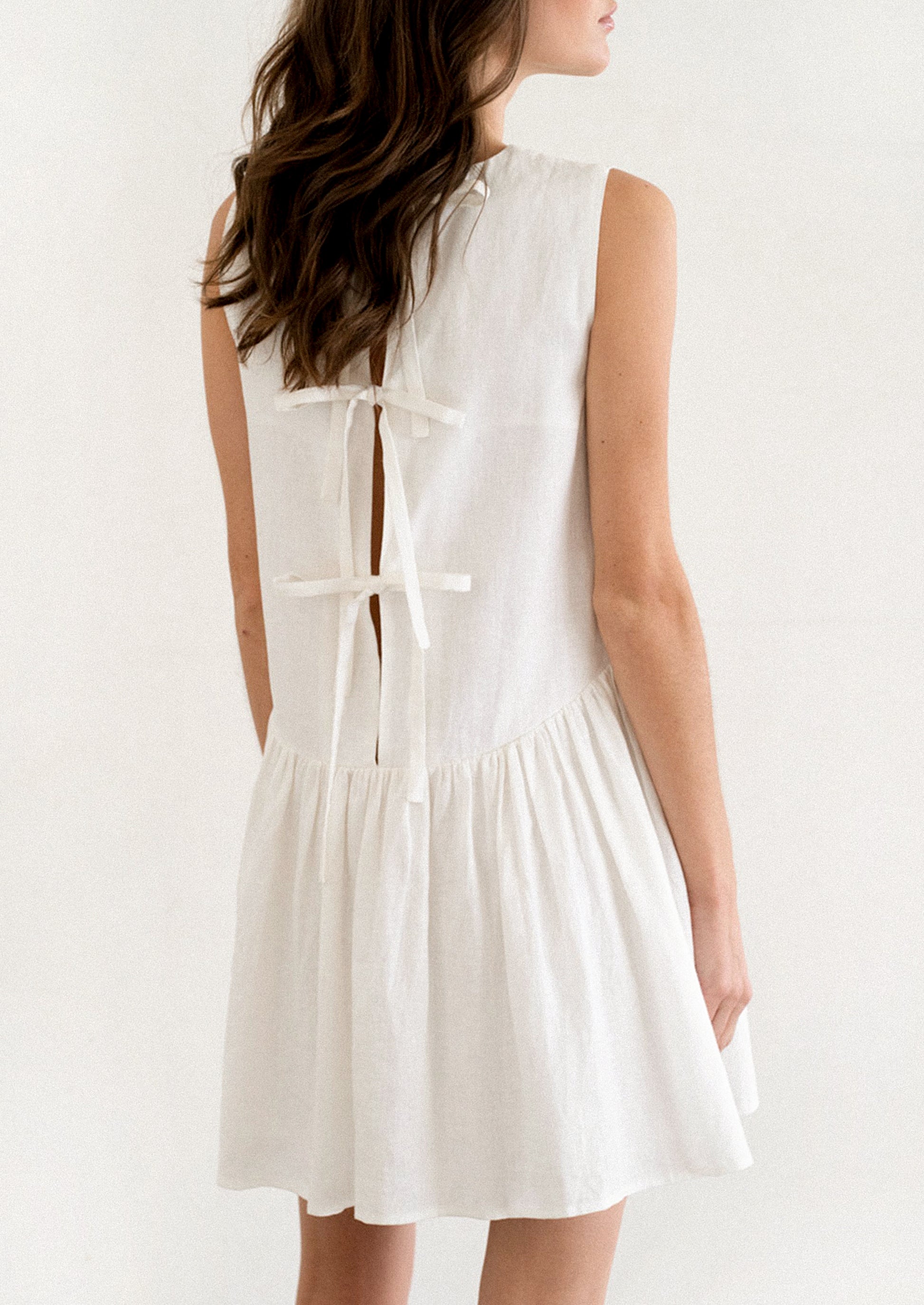 A person wearing a white mini dress with dropwaist and tieback detail.