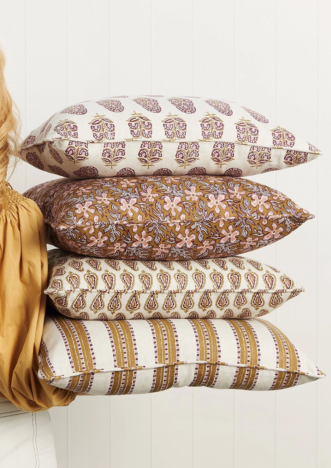 Four decorative throw pillows stacked vertically, held by a person wearing a gold blouse against a white paneled wall. The pillows feature various patterns including paisley, floral, and stripes in earthy tones of brown, beige, and muted pink.