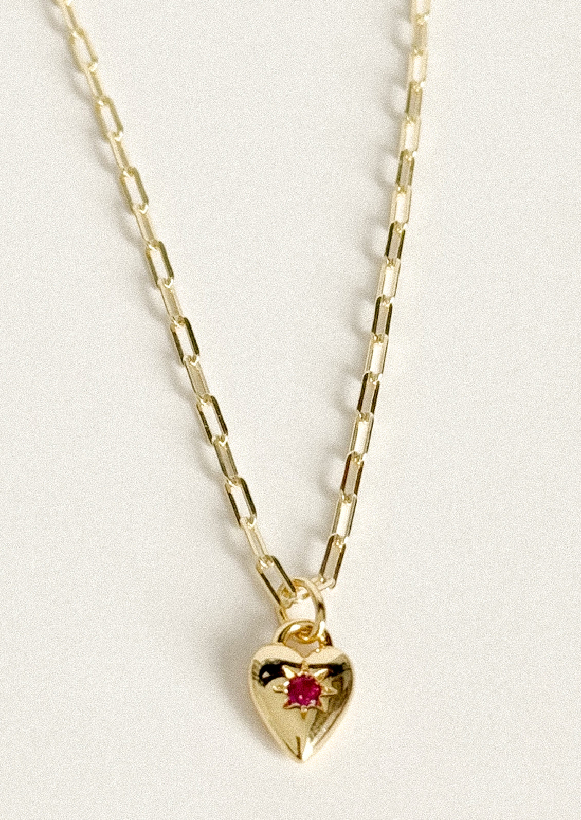 A gold chain necklace with a small heart-shaped pendant featuring a tiny red gemstone in the center, displayed against a light off-white background.