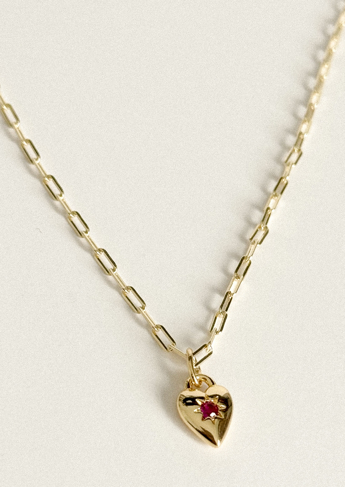 A gold paperclip chain necklace with a small heart-shaped pendant featuring a tiny red gemstone, displayed on a clean off-white background.