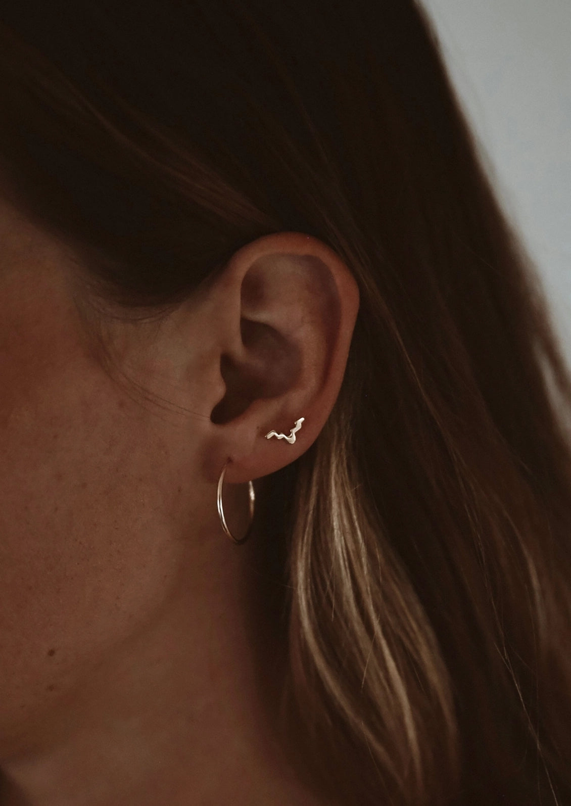 A close-up of a person's ear wearing a delicate silver bat-shaped stud earring and a silver hoop earring, with brown hair in the background.