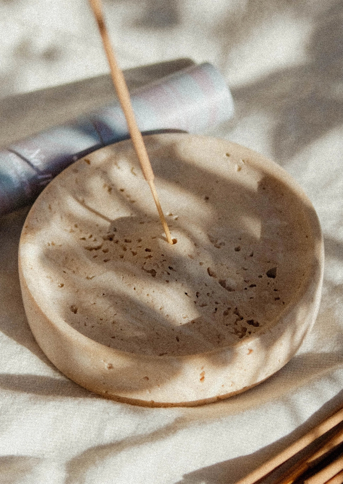 A round beige stone incense holder with a burning incense stick, resting on a textured beige fabric under soft natural shadows.