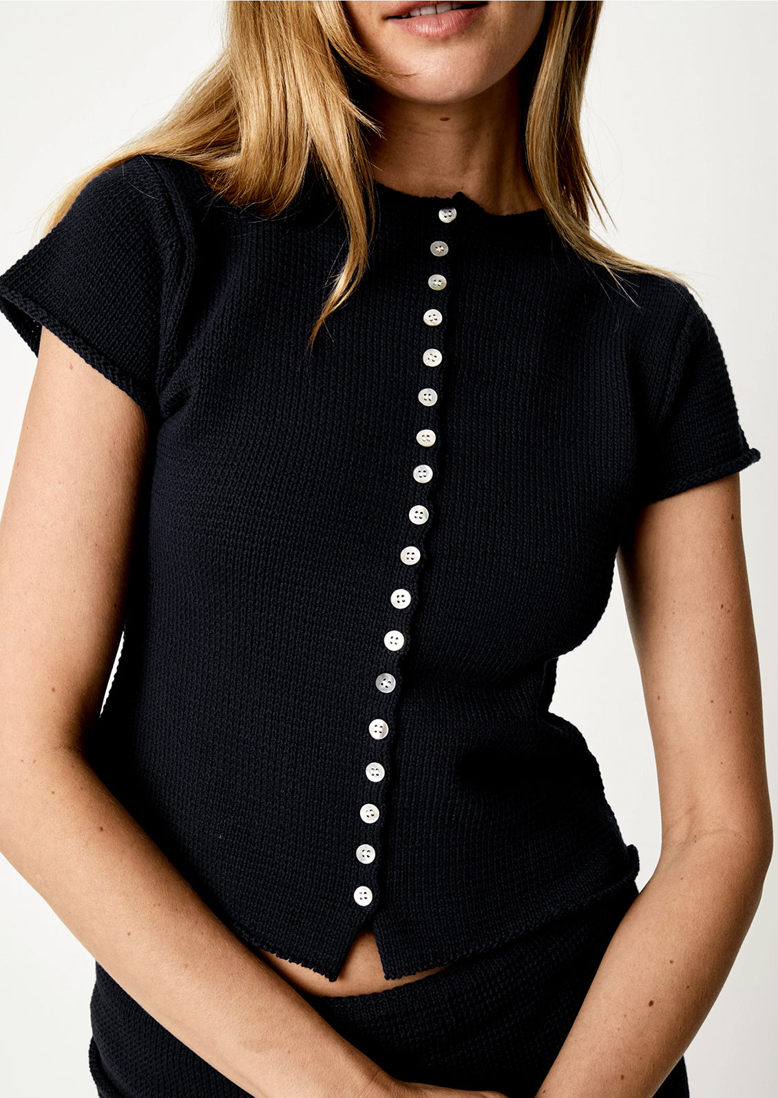 A close-up of a woman wearing a black short-sleeve ribbed knit top with a vertical row of white buttons down the front, against a plain light background.