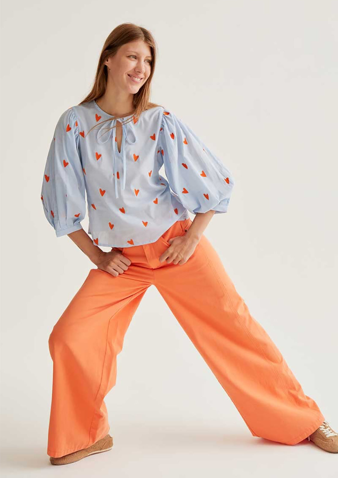 Woman wearing a light blue blouse with red patterns and orange pants on a white background