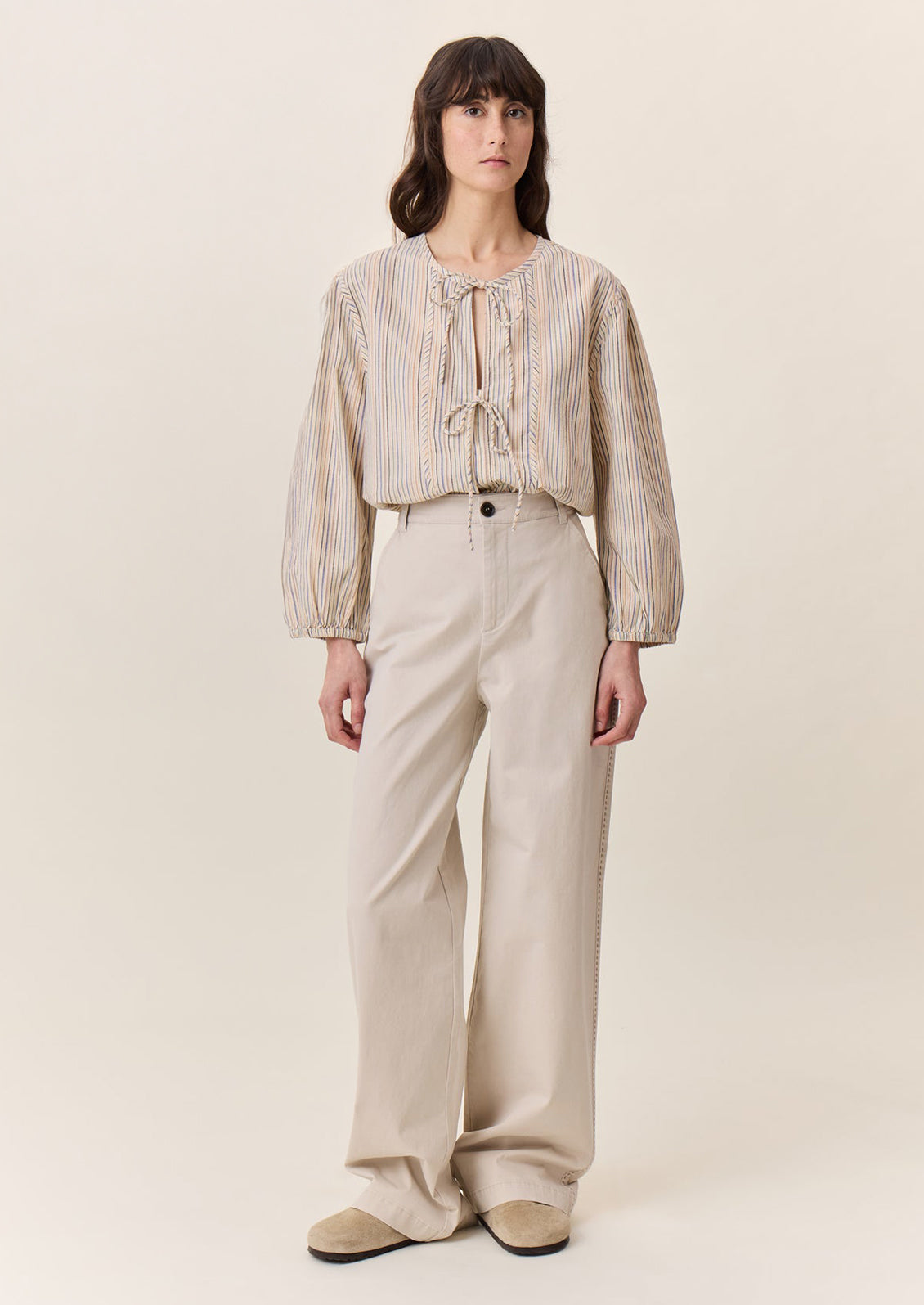 A woman wearing a tie neck blouse in cream with pastel multicolor stripe pattern.