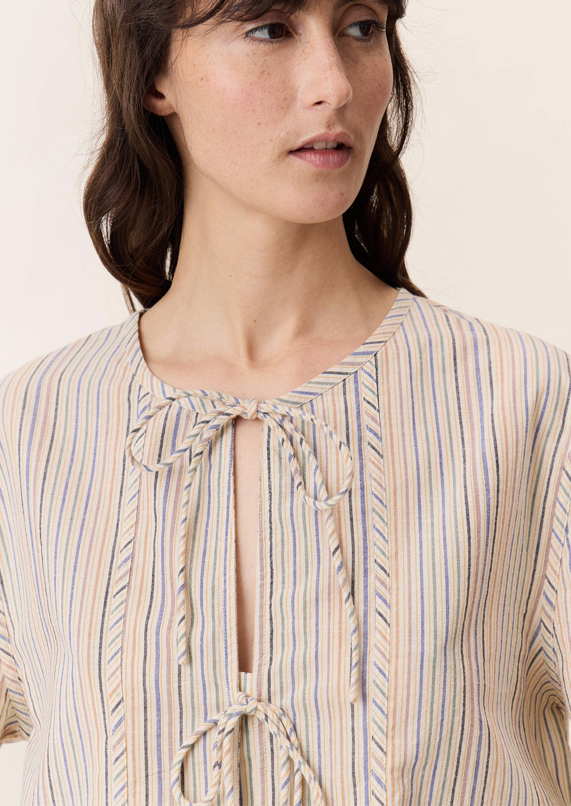 A woman wearing a tie neck blouse in cream with pastel multicolor stripe pattern.