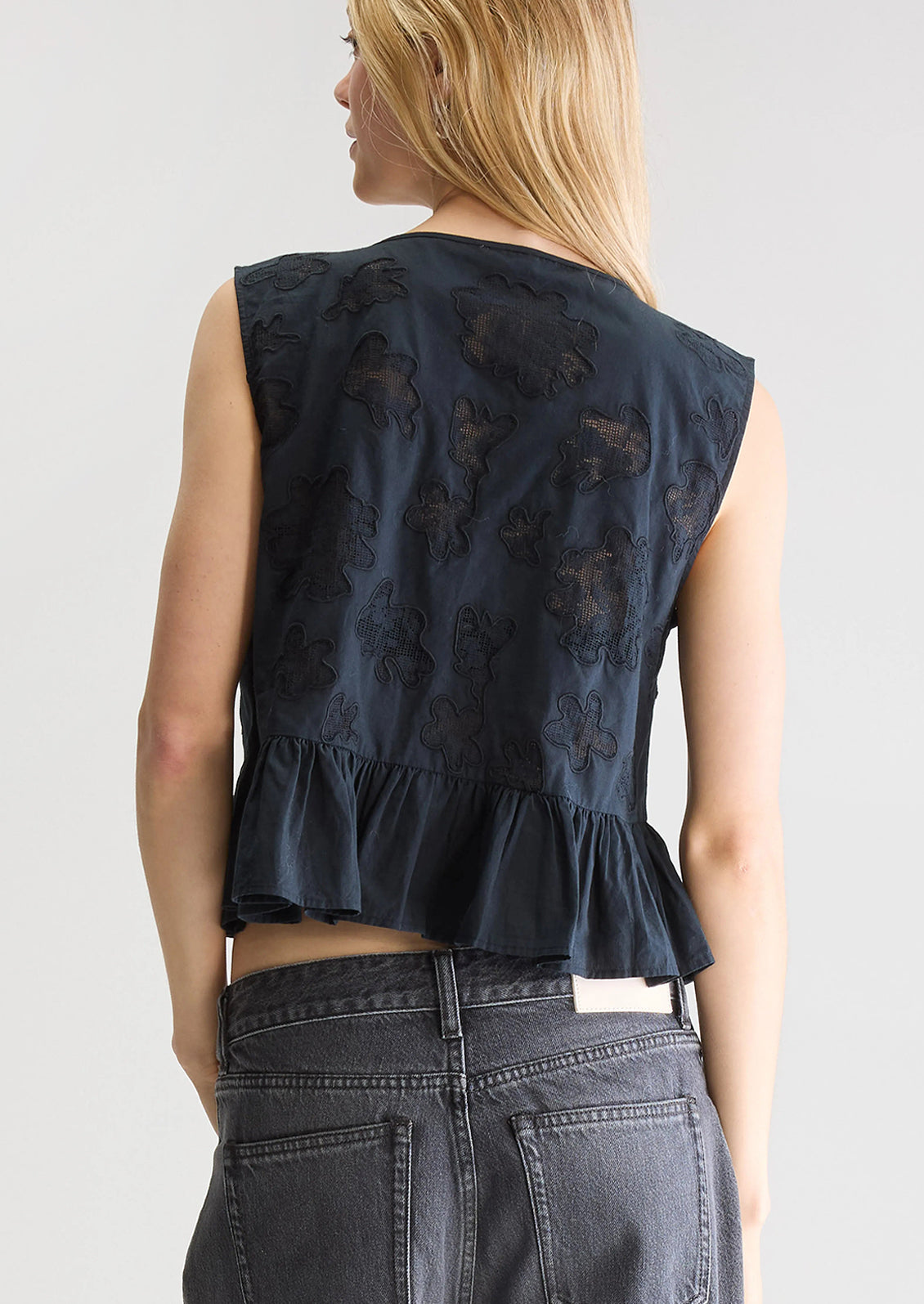 A woman wearing a sleeveless tie front top in black with tonal lacework detailing and peplum hem.