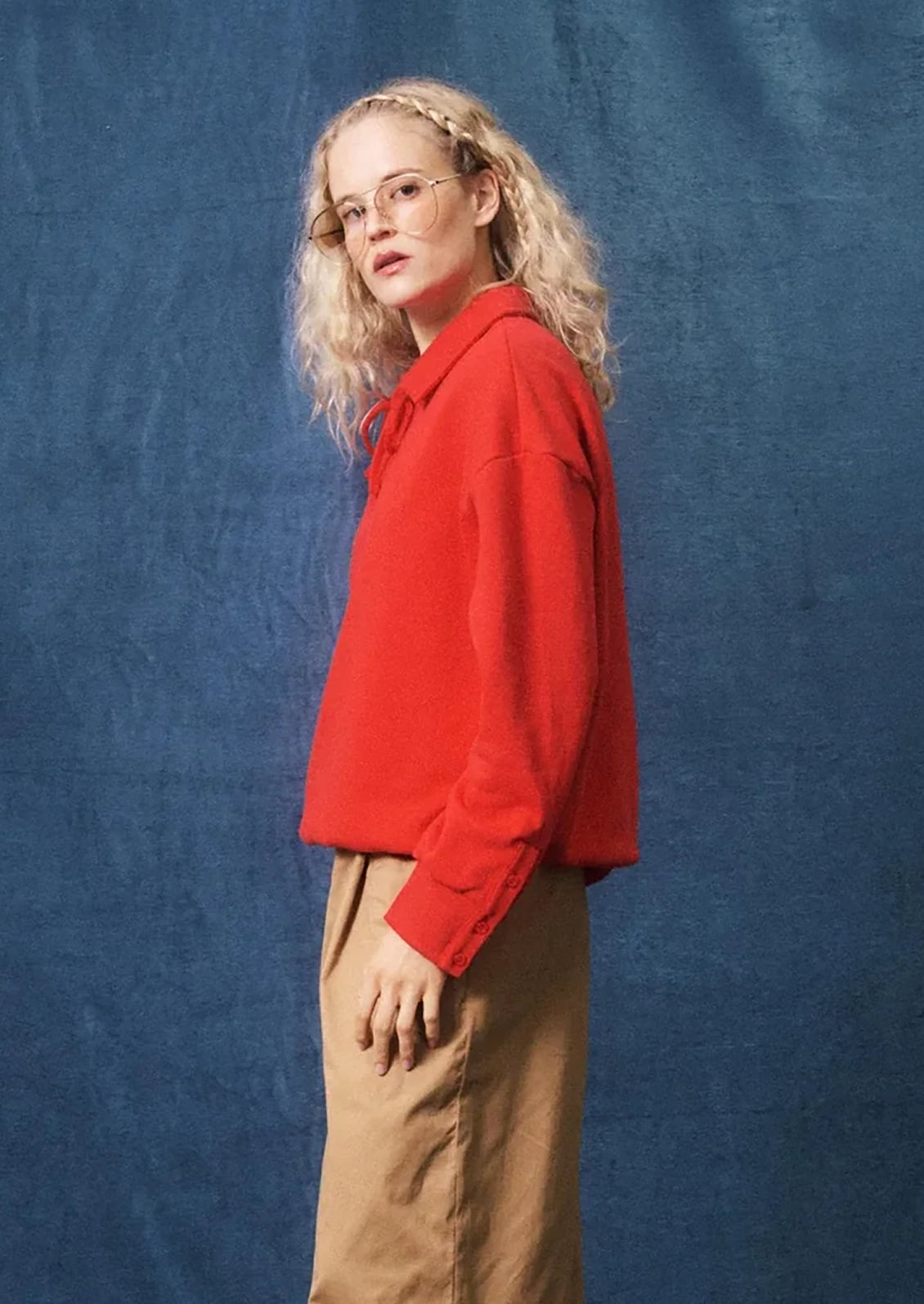 A woman with blonde braided hair and glasses, wearing a bright red long-sleeve cropped pullover and tan pants, standing against a textured blue backdrop.