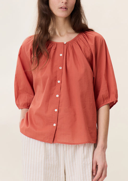 A woman wearing a short sleeve blouse in coral red color with shirring at neckline.