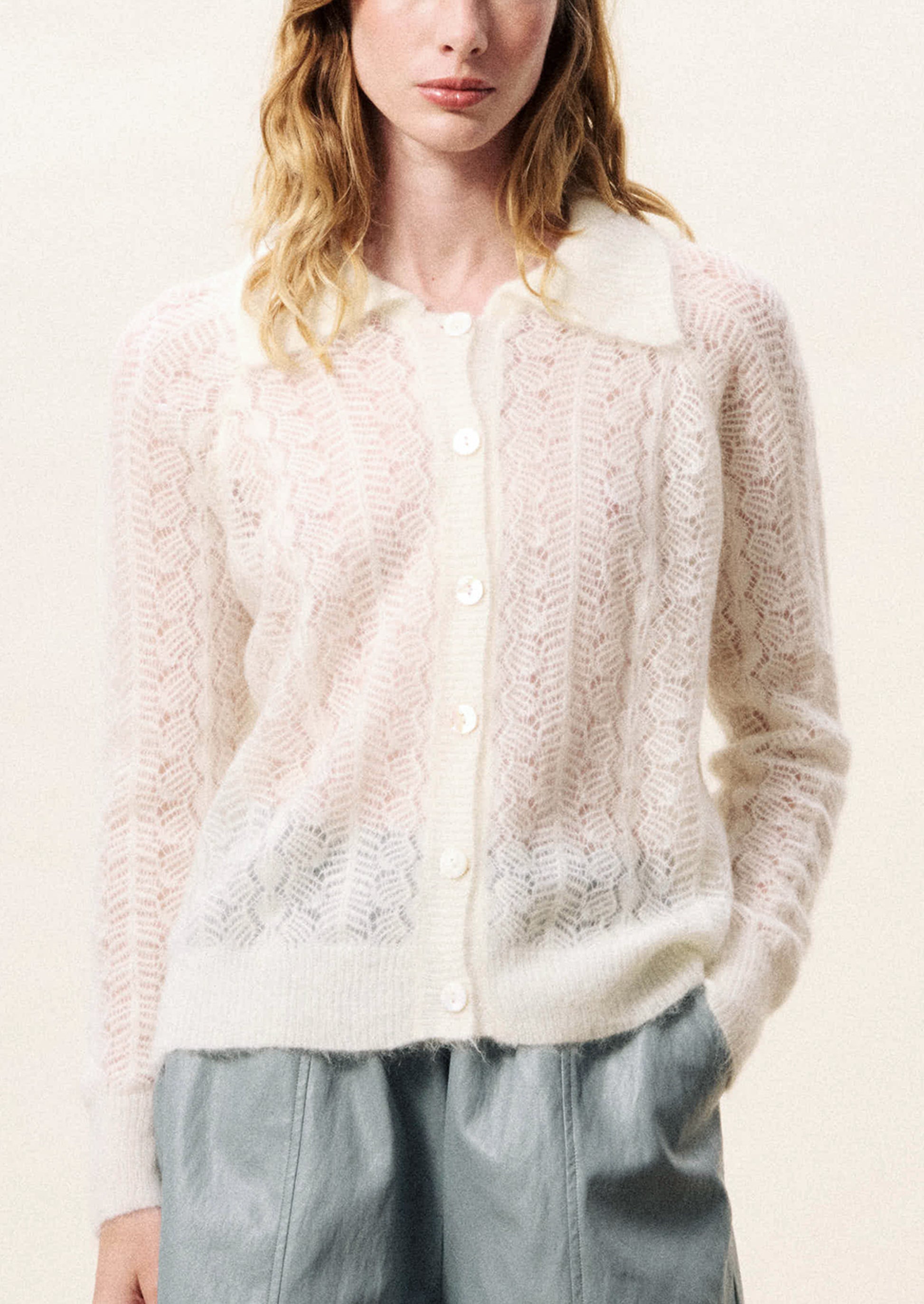 Person wearing a white lace cardigan with arms crossed on a light background