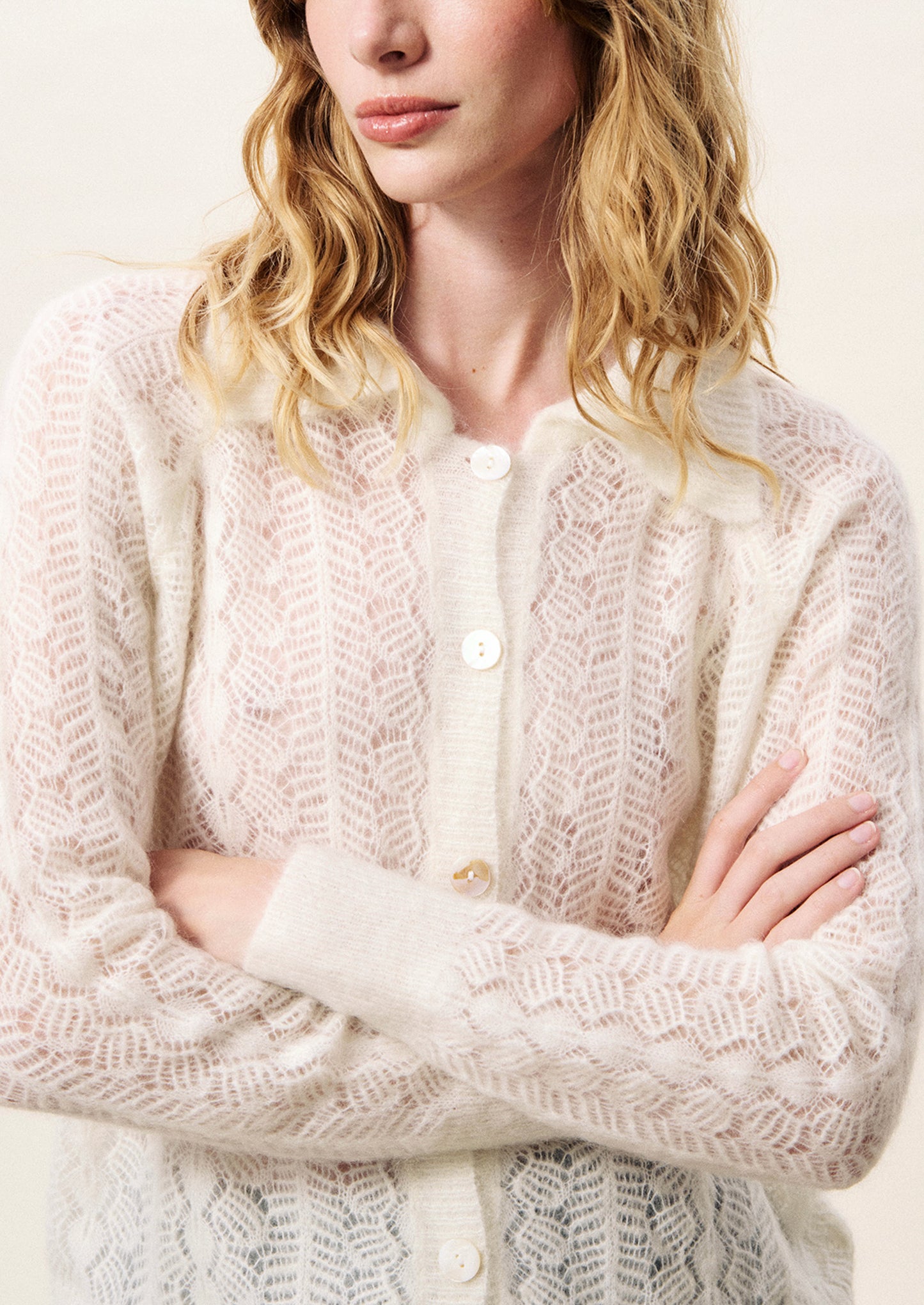 Person wearing a white lace cardigan with arms crossed on a light background
