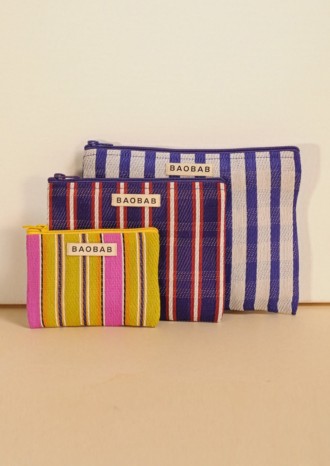 Three striped fabric pouches from BAOBAB, arranged by size against a plain beige background. The smallest pouch is yellow, pink, and orange striped; the middle pouch is navy, red, and white striped; the largest pouch is blue and white striped.