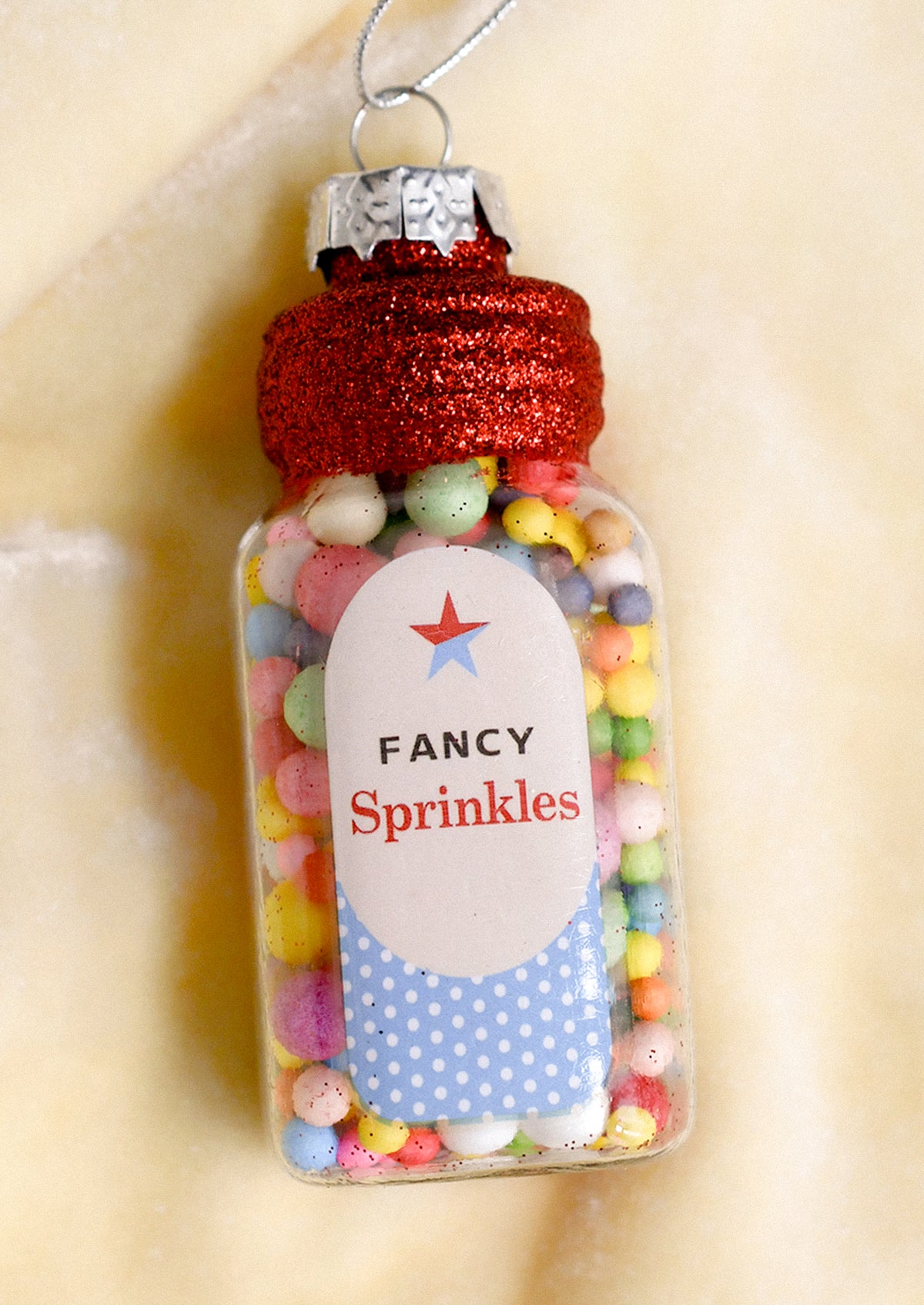 A clear glass bottle ornament filled with pastel multicolor sprinkles, topped with a glittery red cap and a hanging silver wire. The front label reads 'Fancy Sprinkles' with a red star and blue polka dot details.