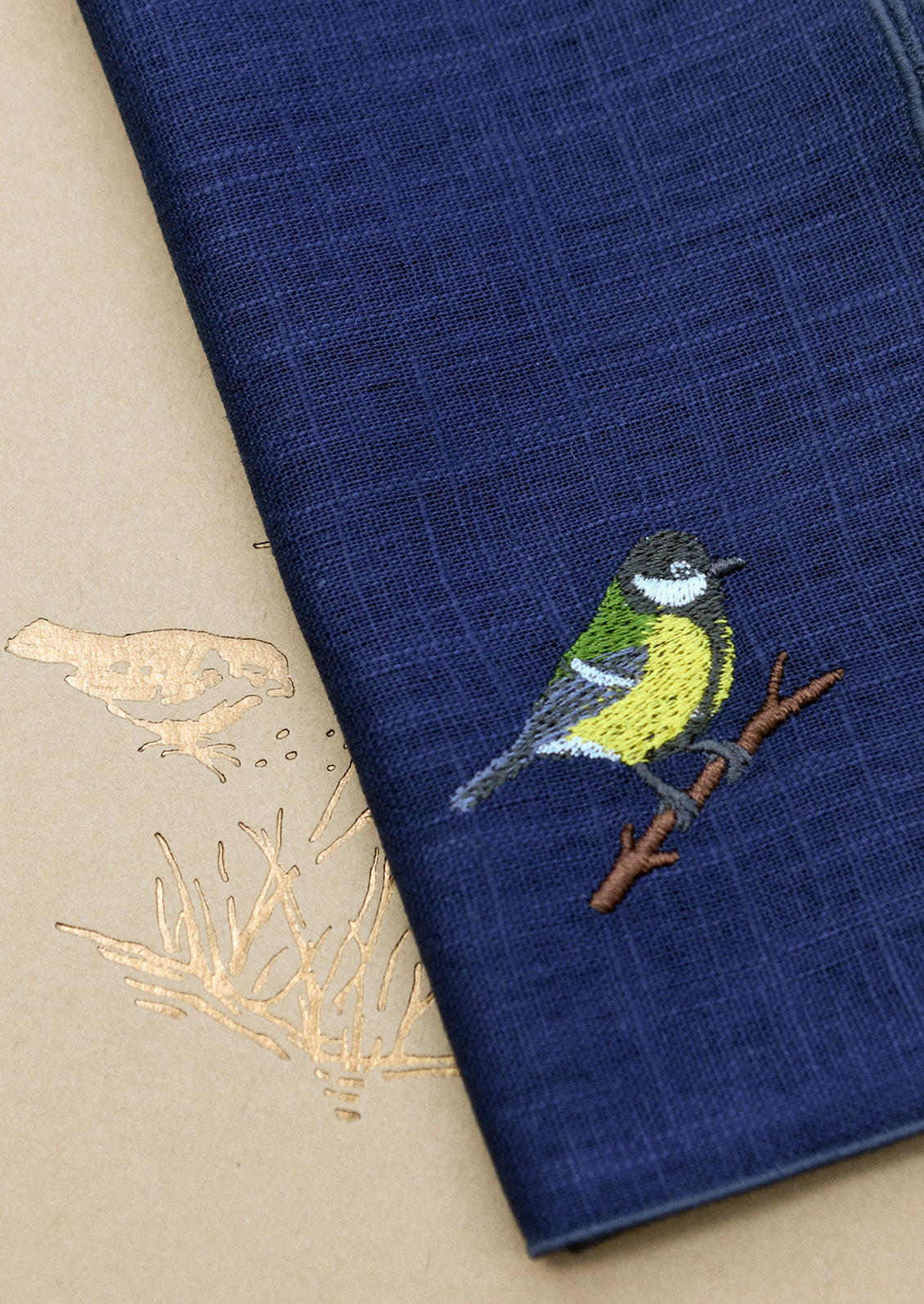A navy blue linen napkin with tonal serged edges, and an embroidered bird at corner.