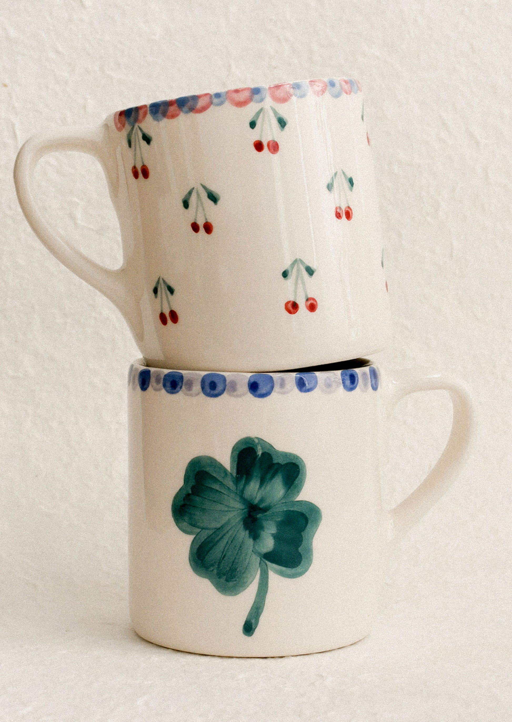 A pair of two mugs, one with painted cherry pattern, one with painted clover.
