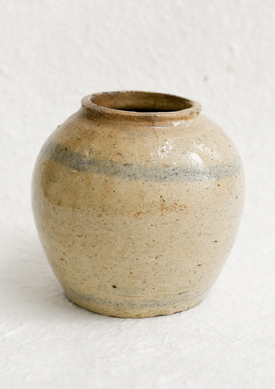 A small antique ginger jar in distressed tan ceramic with pale blue stripes.