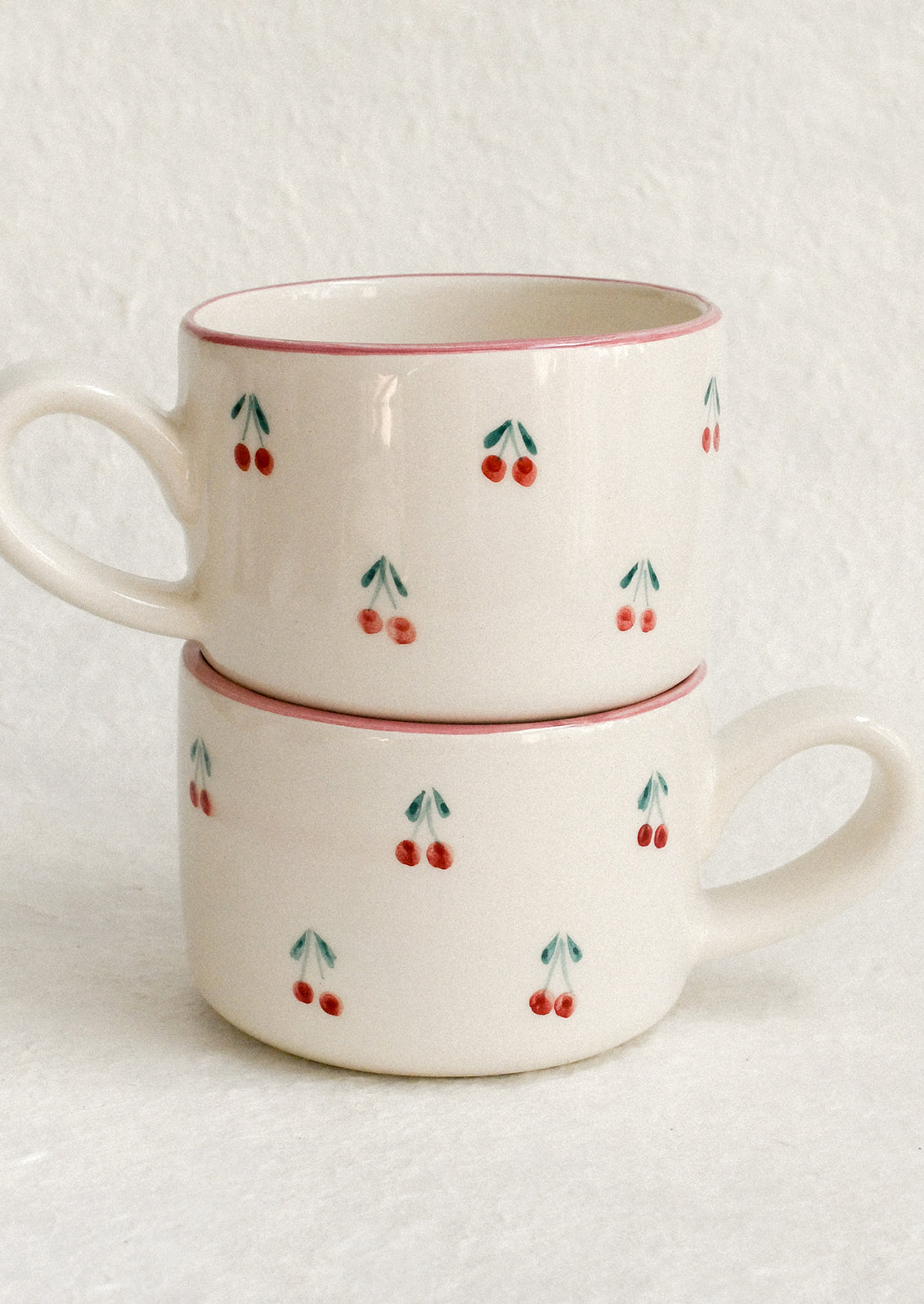 A cream ceramic mug with hand painted cherry motif and pink top rim.